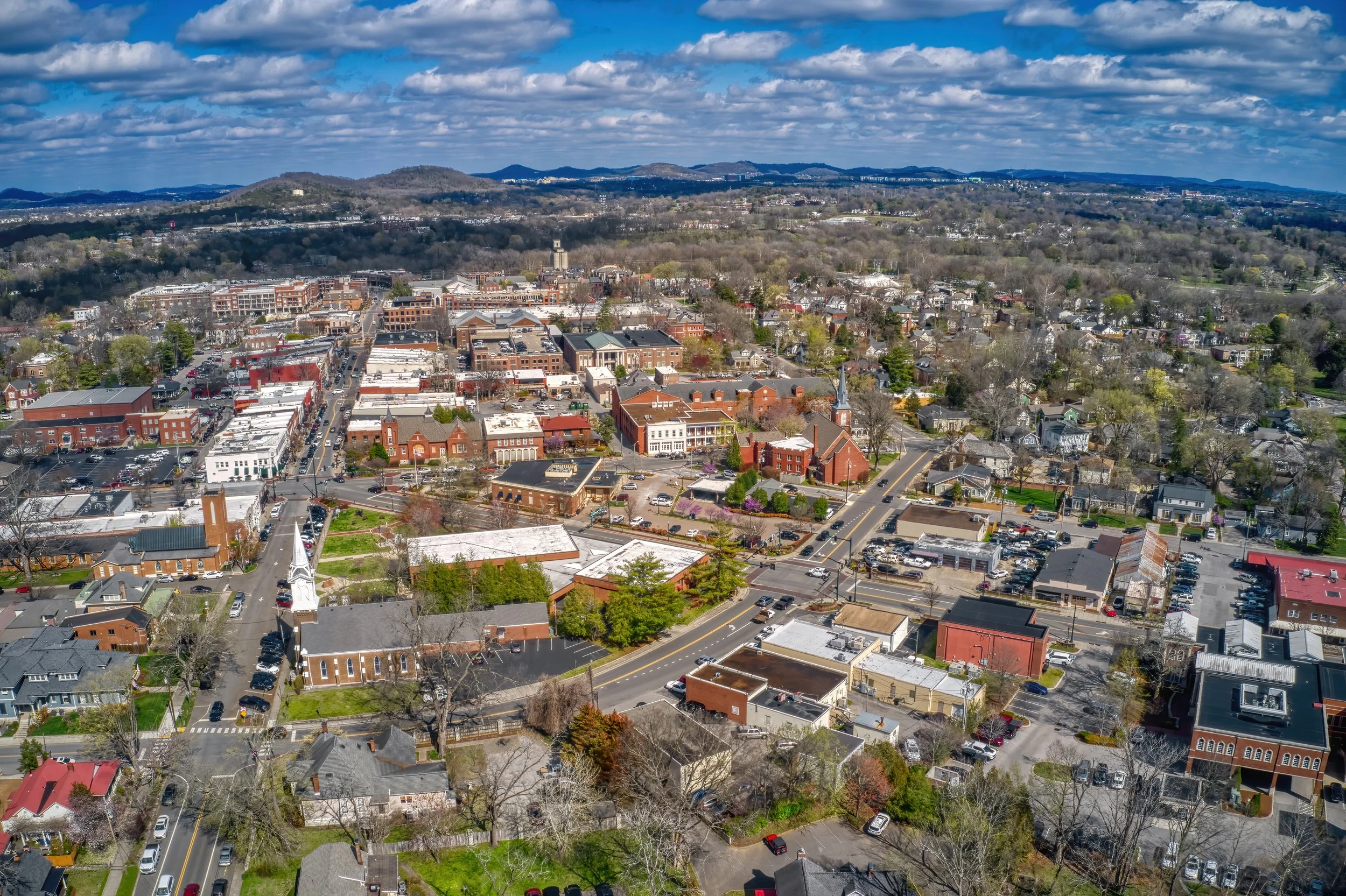 Aerial view of a small town with streets, buildings, and trees, surrounded by hills and mountains under partly cloudy sky.