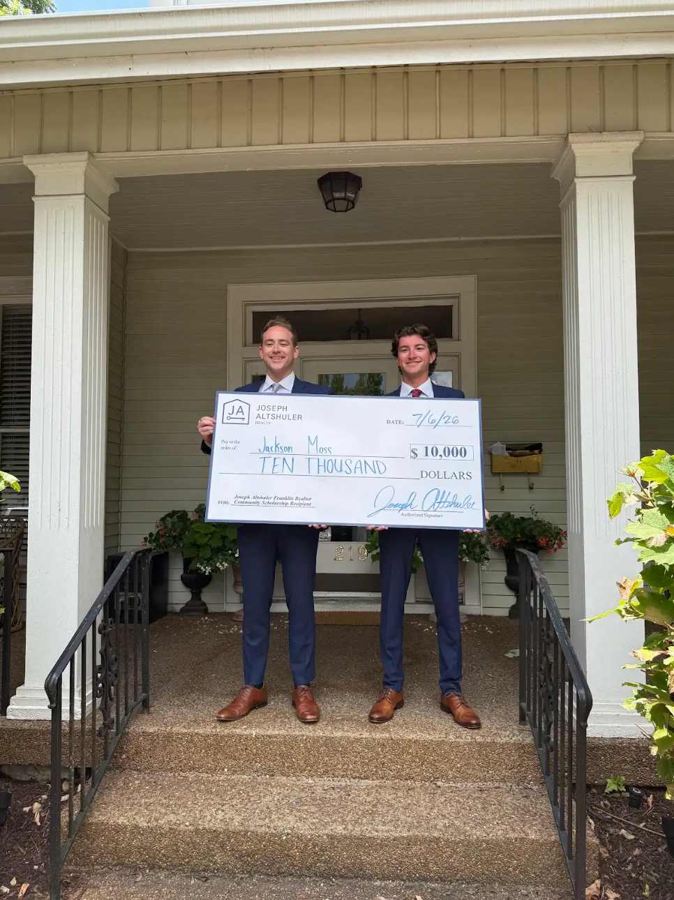 Two young men in suits standing on the porch of a house, holding an oversized check for $10,000 made out to Jackson Moss, with both men smiling.
