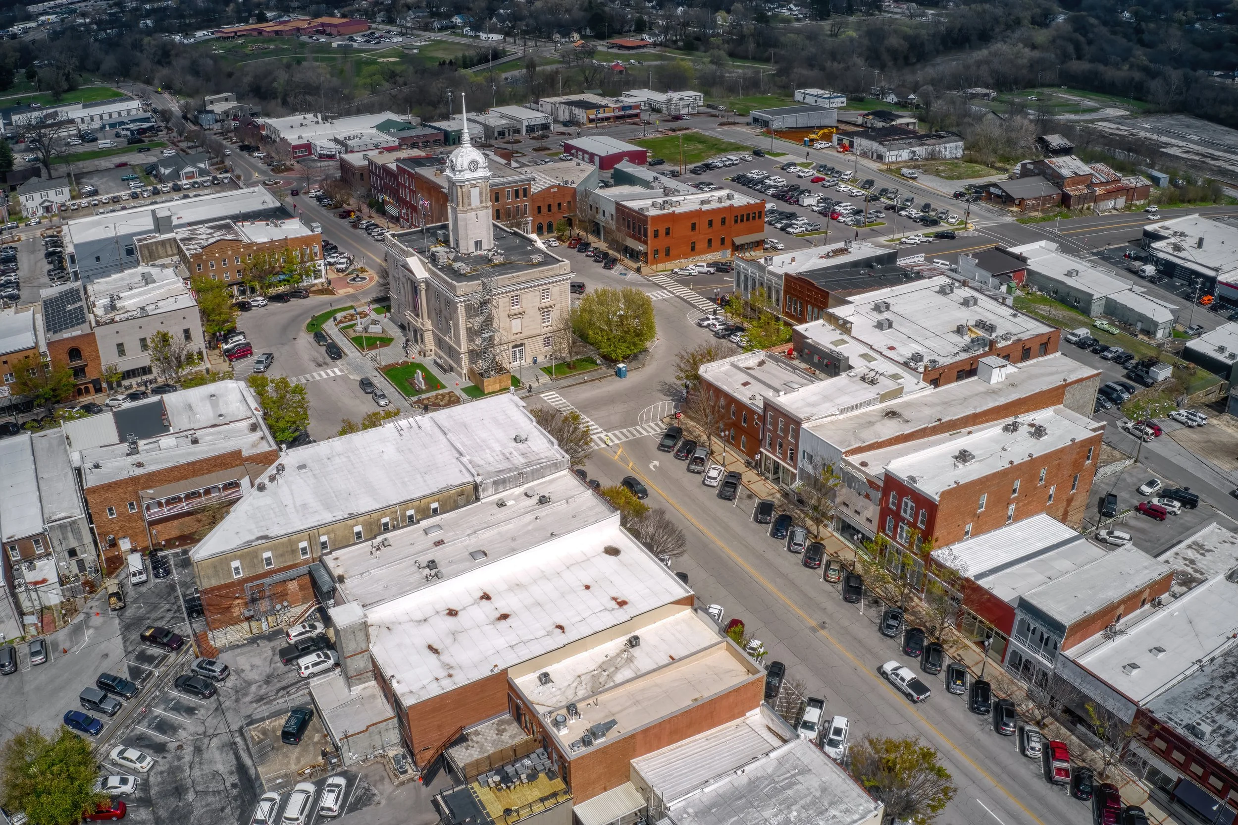 Aerial view of a small town downtown area with a clock tower, surrounding older brick buildings, and a parking lot filled with cars, with trees and green spaces in the background.