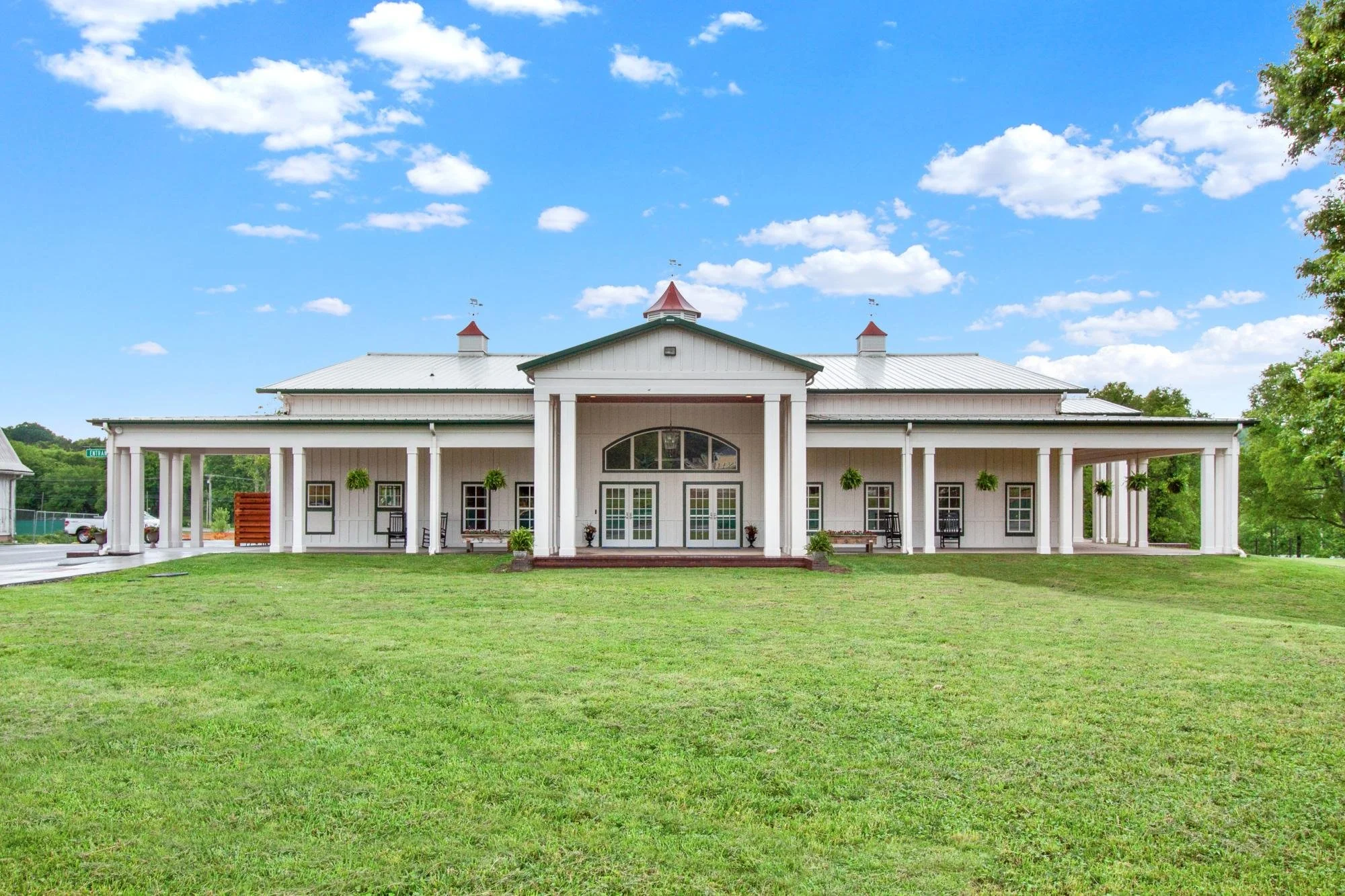 A large, white, two-story house with a metal roof, surrounded by a green lawn and tall trees under a blue sky with scattered clouds.