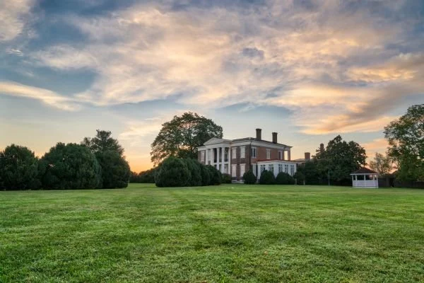 A large, historic house surrounded by lush green grass and trees at sunset, with a partly cloudy sky.