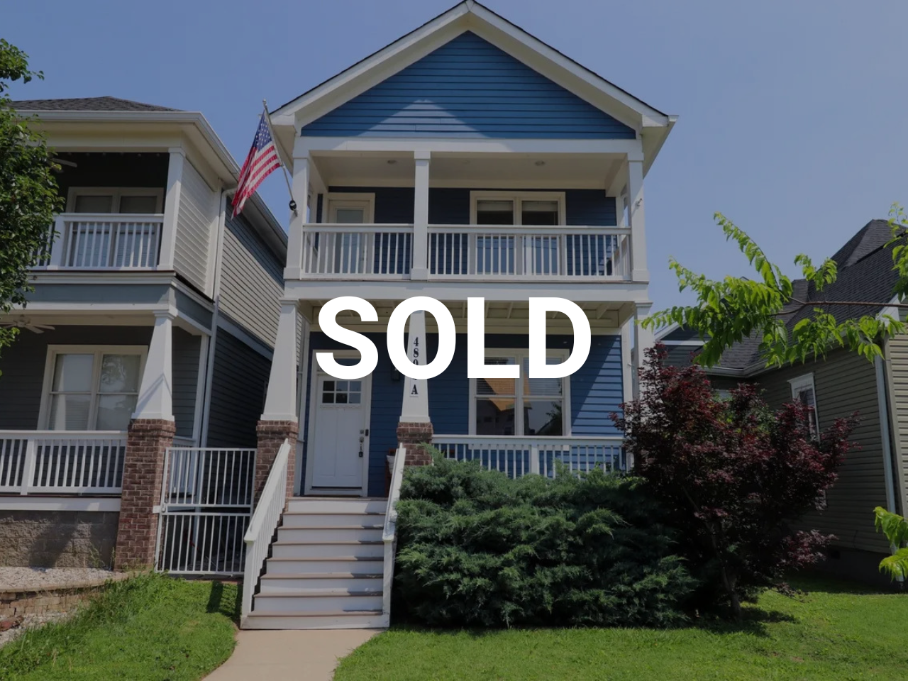 Blue two-story house with white trim, front porch, metal railing, American flag, surrounded by green bushes and trees, with a clear blue sky, 'SOLD' overlay.