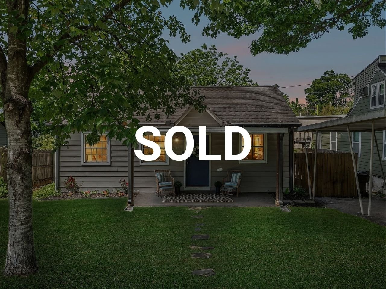 A house with two windows lit from inside, a front porch with outdoor seating, and a large tree in the front yard. The word 'SOLD' is overlaid on the image.