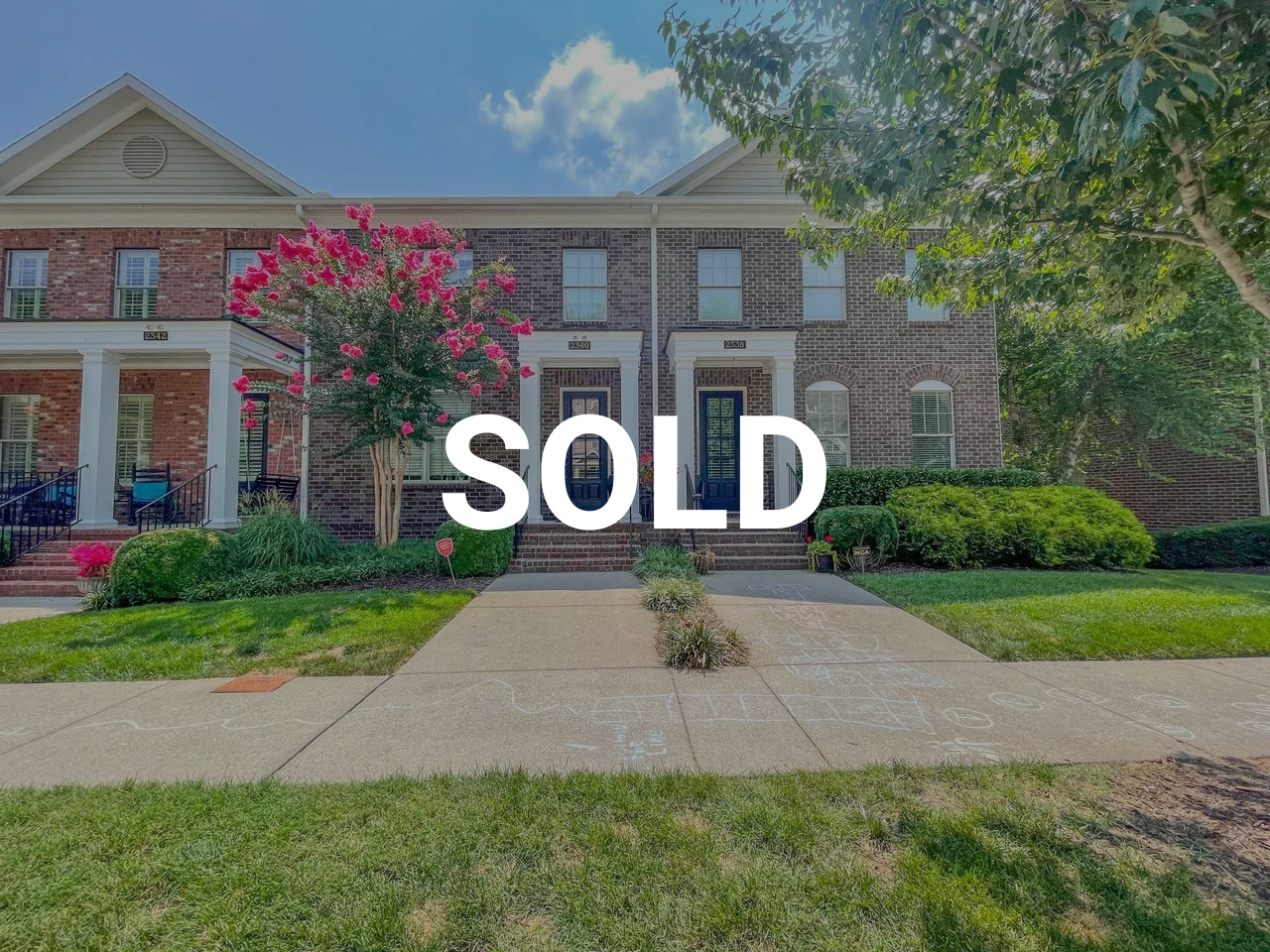 A residential brick house with a front porch and a well-maintained lawn. There are trees and shrubs, with a painted hopscotch game on the sidewalk. The word 'SOLD' is overlaid on the image.