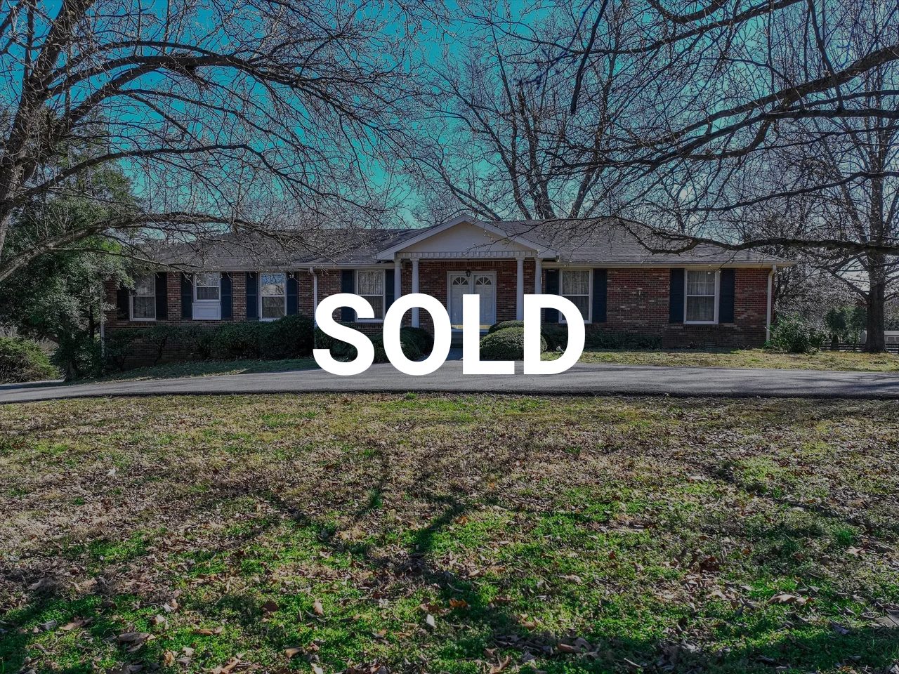 A single-story brick house with a white front door and windows, surrounded by leafless trees and a grassy yard with fallen leaves, with a clear blue sky above.