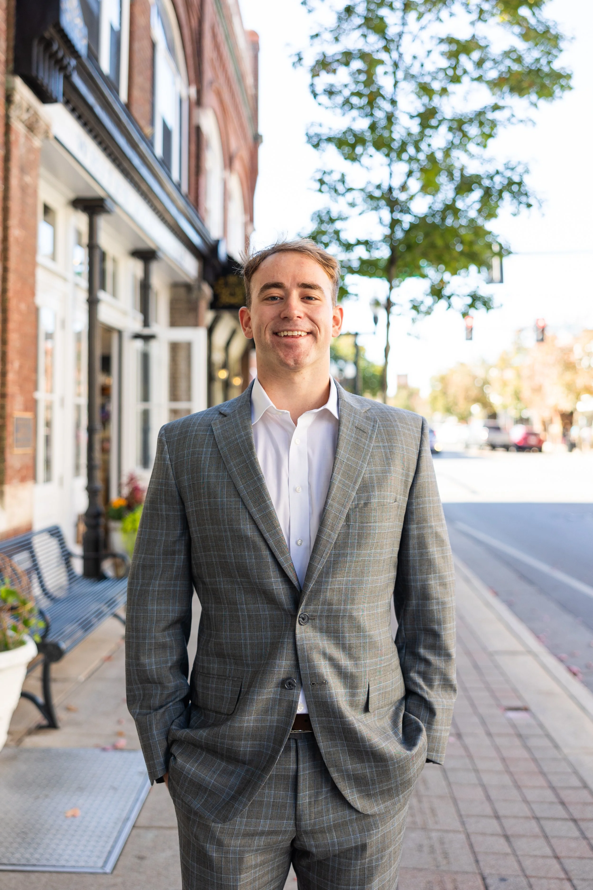 A young man in a gray plaid suit smiling on a city sidewalk.