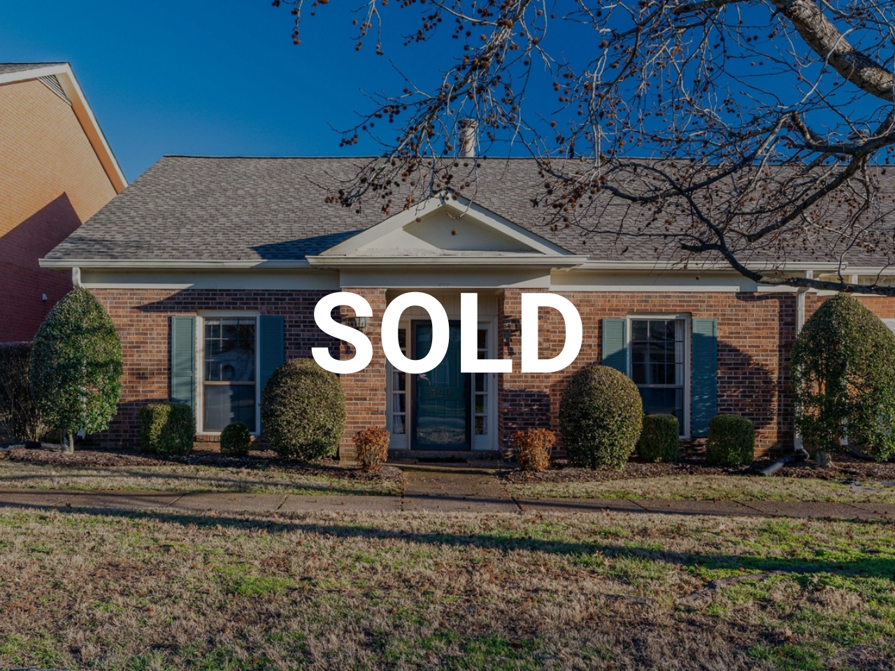 Front view of a brick house with four windows, bushes, and a tree with no leaves in front, under a blue sky, with a 'SOLD' overlay.