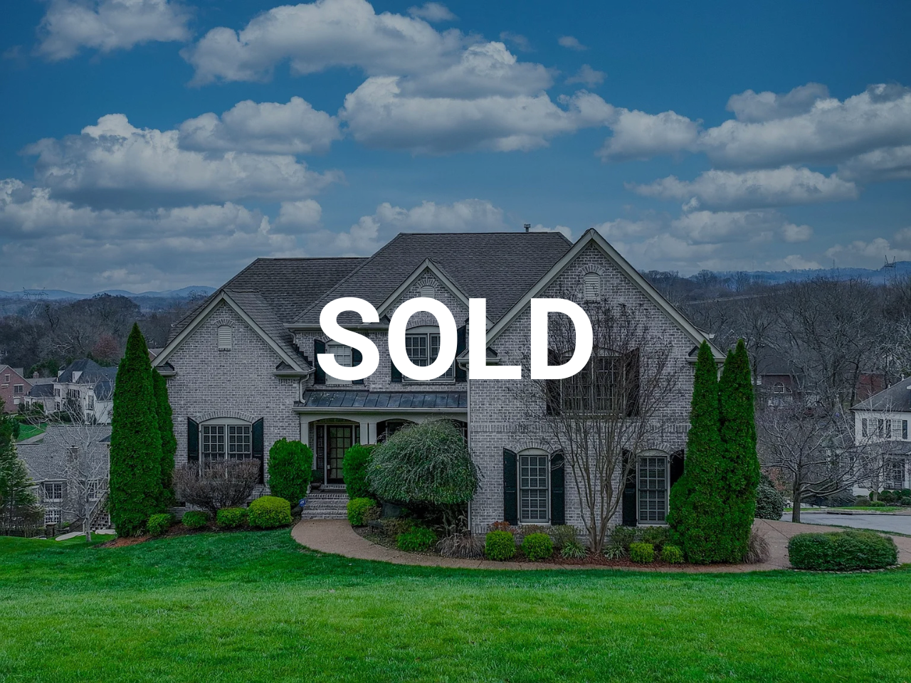 Front view of a two-story brick house with a well-maintained lawn, trees, and shrubbery, under a partly cloudy sky, with the word "SOLD" overlaid.