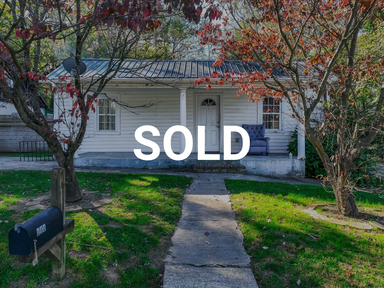A white house with a metal roof, a small front porch with a blue couch, and red-leaved trees in the yard, with a black mailbox labeled '219' in the foreground. The word 'SOLD' is overlaid in large letters.