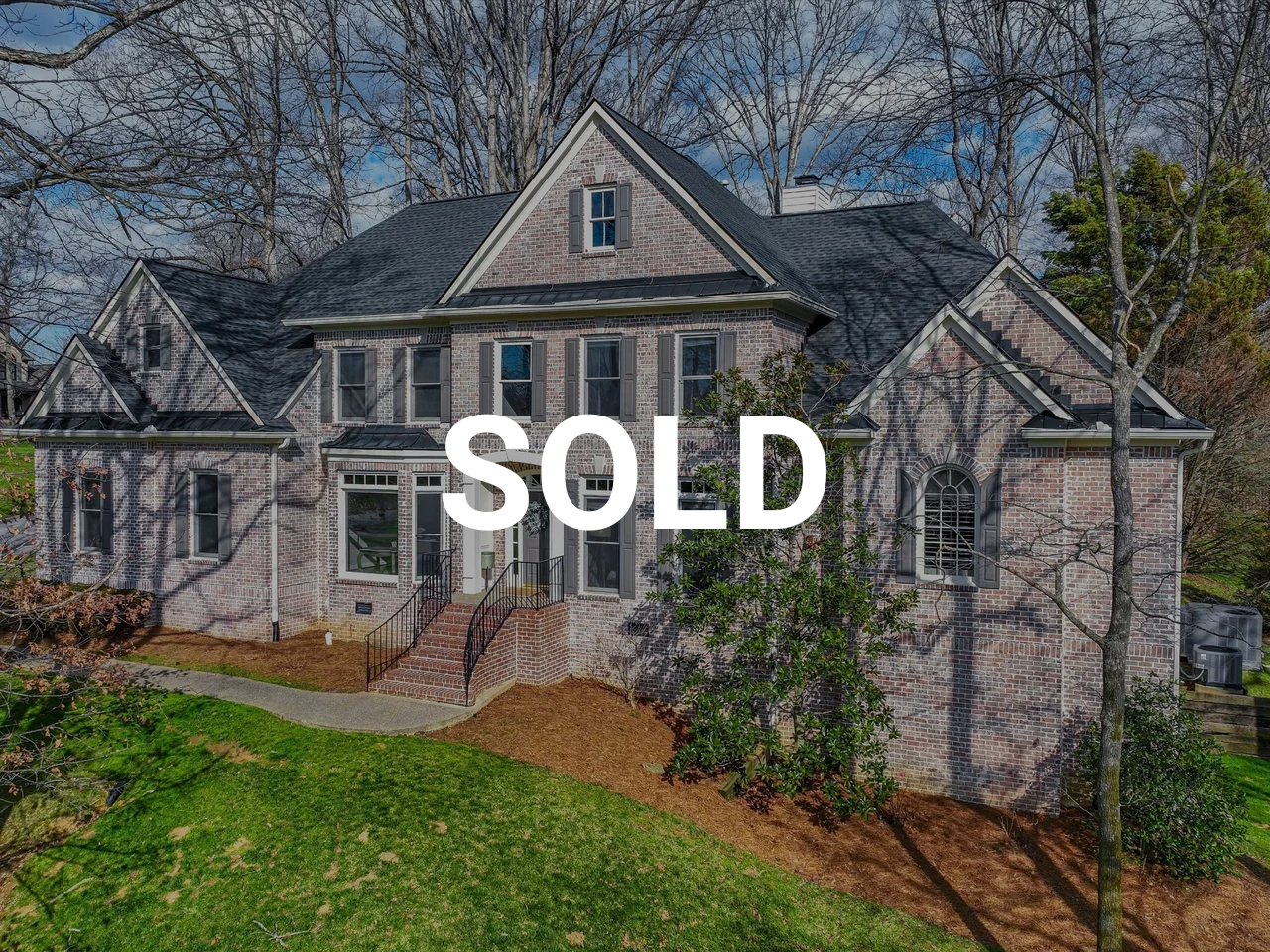 A large brick house with multiple gables, dark roof, and front steps, surrounded by trees and a lawn, with a 'SOLD' sign overlaid.