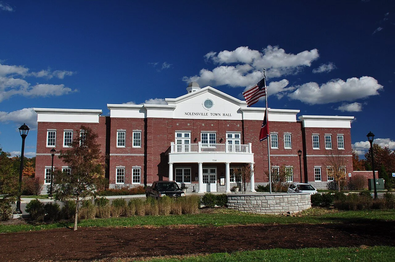 Front view of Nolensville Town Hall, a red brick building with white accents, two flagpoles with American flags in front, under a partly cloudy blue sky.