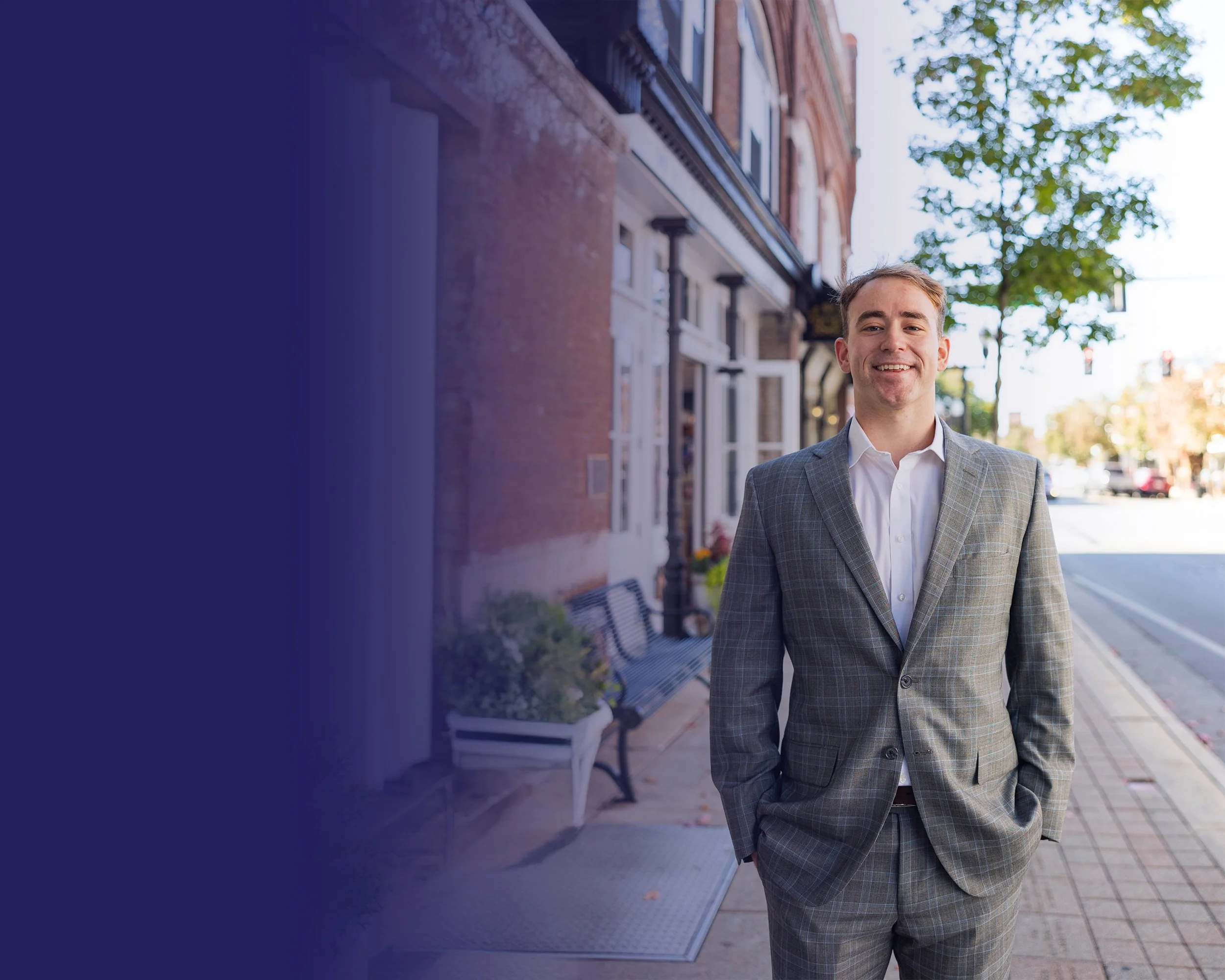 A young man in a plaid gray suit with hands in pockets, smiling, standing on a city sidewalk with trees, brick buildings, and a bench in the background.