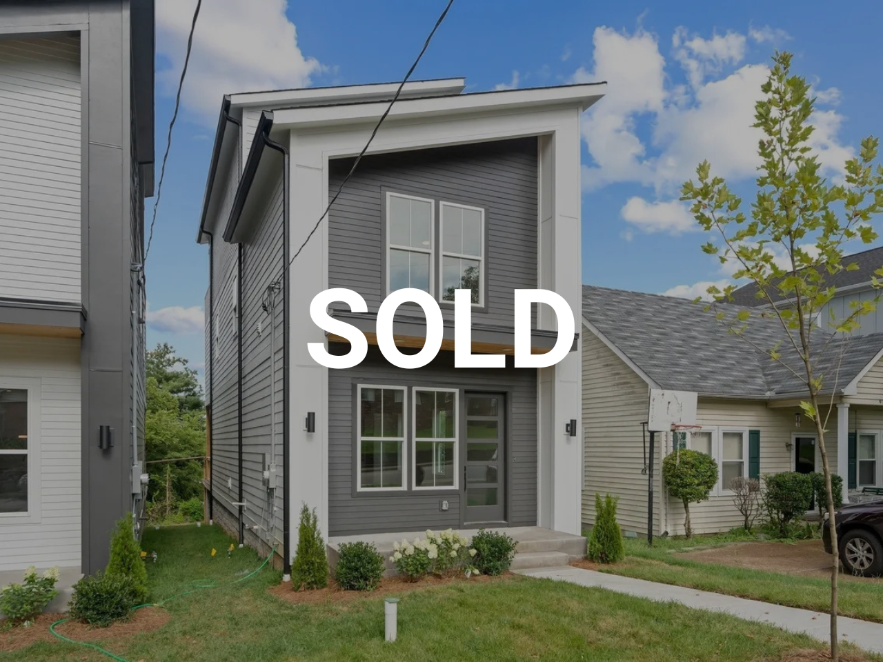 Modern gray house with white trim, front stairs, and large windows, with a lawn and trees in a neighborhood, marked as sold.