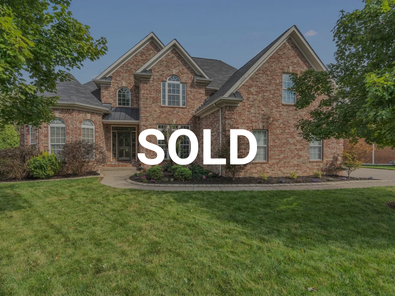 Brick house with a well-maintained lawn, trees, and garden. The house has multiple gabled roofs and arched windows. The word "SOLD" is overlaid on the image.