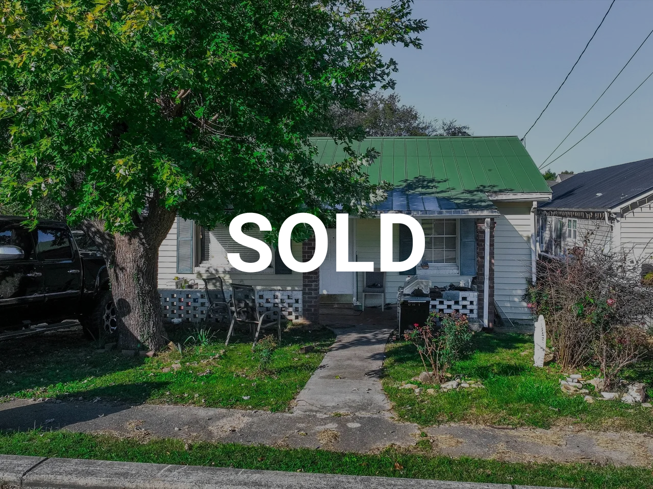 Front view of a small house with a green metal roof, large tree on the left, outdoor chairs and a porch, and a