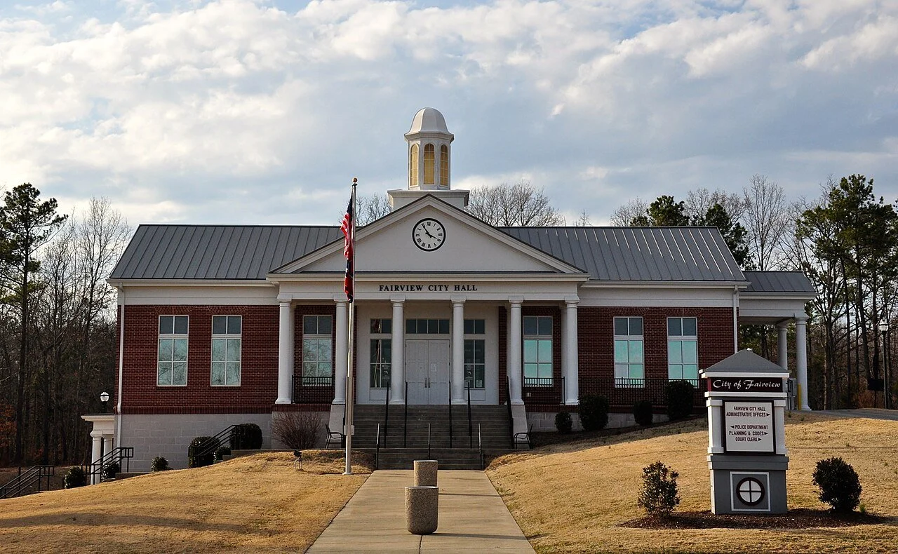 Front view of Fairview City Hall building with a clock, American flag, and a sign for the city in a grassy area with trees in the background.