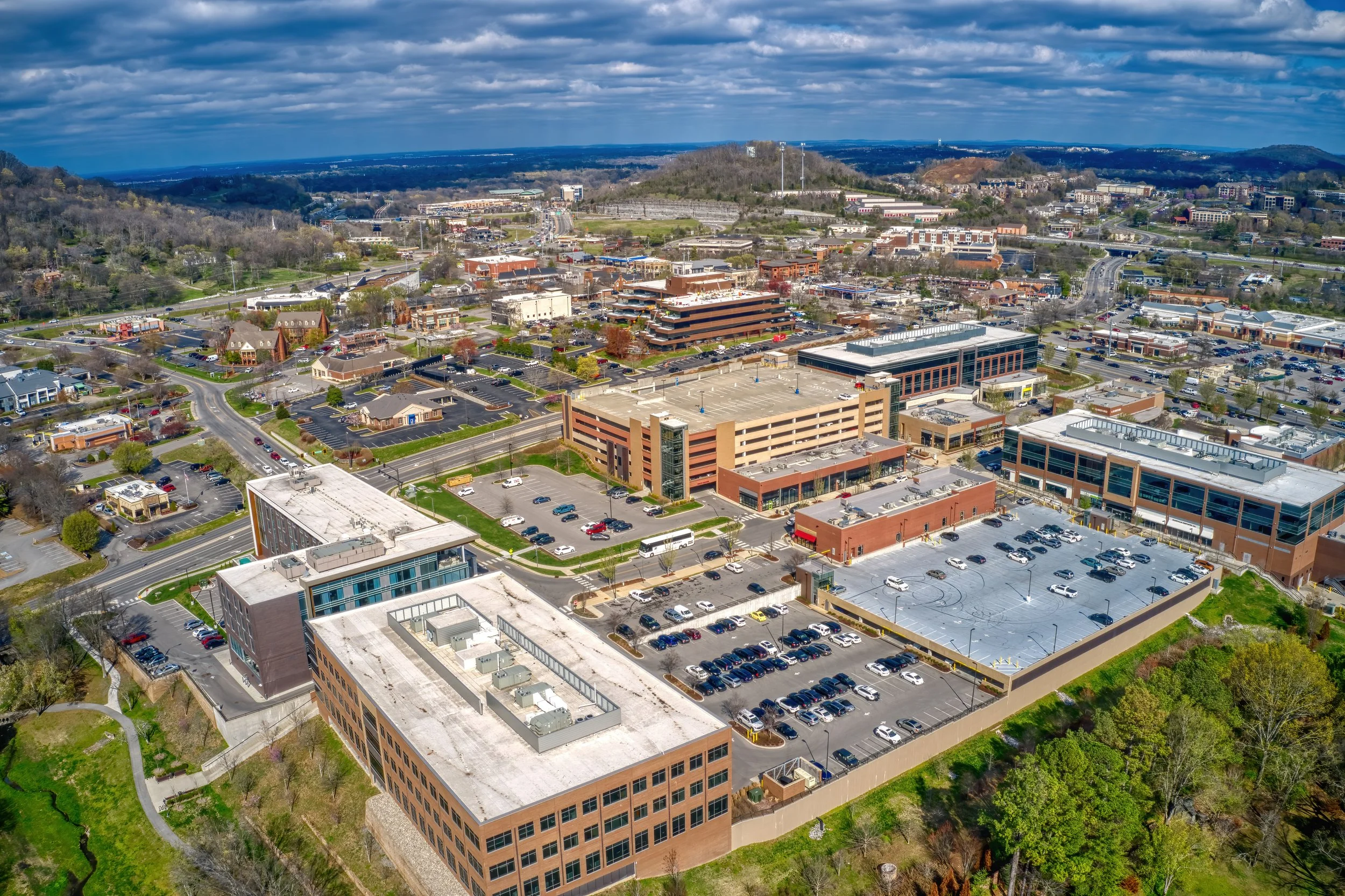 Aerial view of a commercial area with parking lots, office buildings, and surrounding greenery, with hills and a cloudy sky in the background.