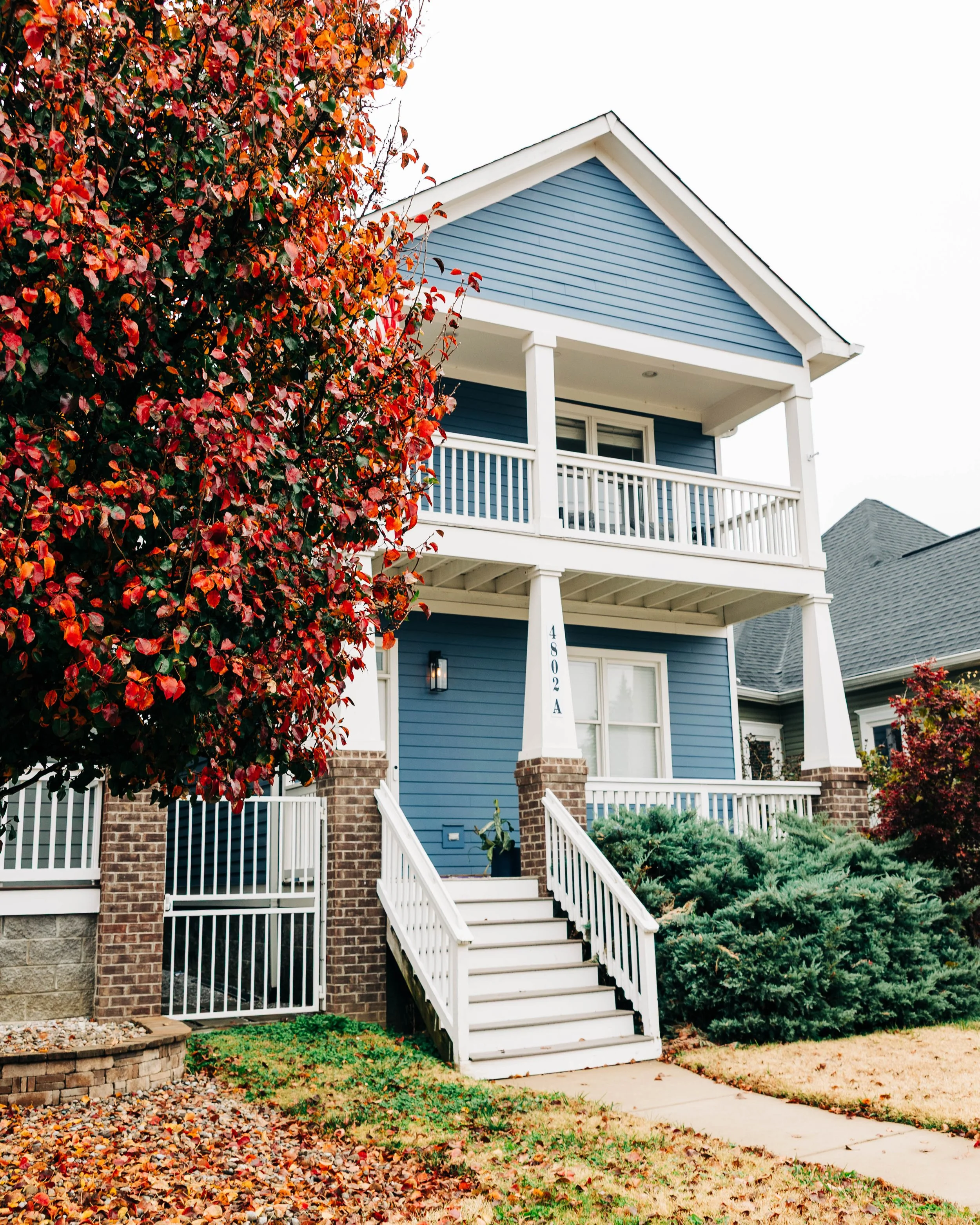 A two-story blue house with white railings, stairs, and trim, surrounded by autumn foliage and shrubs.