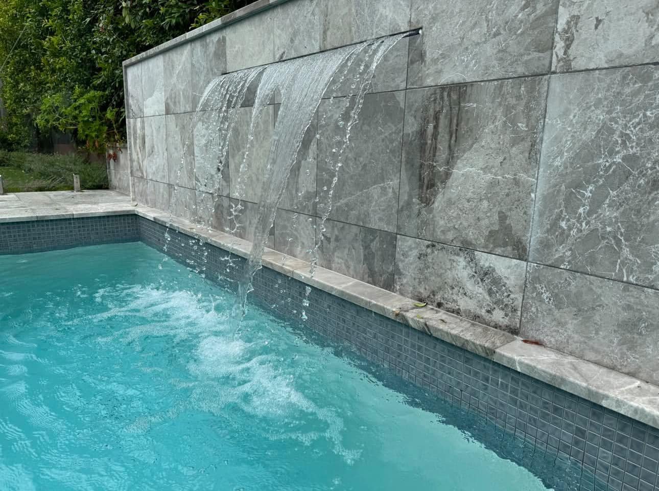 A swimming pool with three water fountains flowing into it, set against a stone wall with greenery in the background.