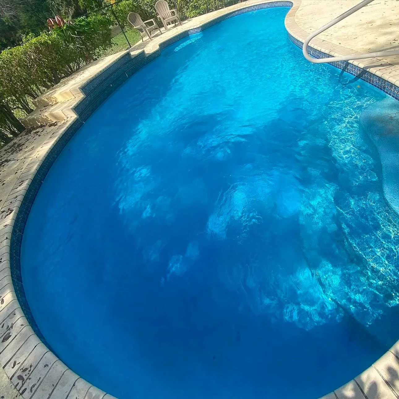 An outdoor swimming pool with blue water, surrounded by a concrete deck, with patio chairs and greenery in the background.