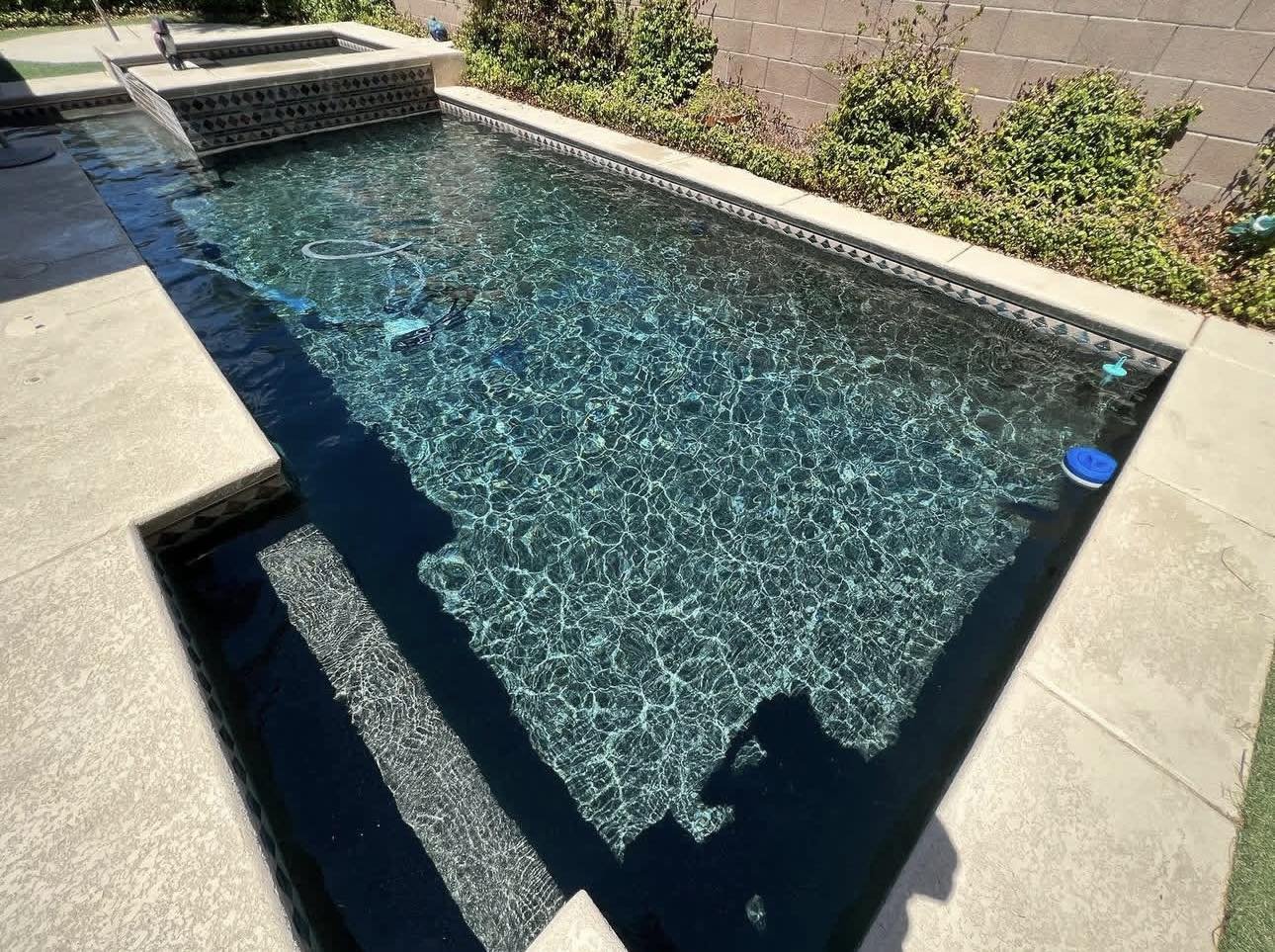 Photo of a rectangular in-ground swimming pool with a small waterfall feature on one corner. The pool has clean, clear water with visible ripples and a textured bottom. There are beige concrete pool surrounds and a brown brick wall with green bushes 