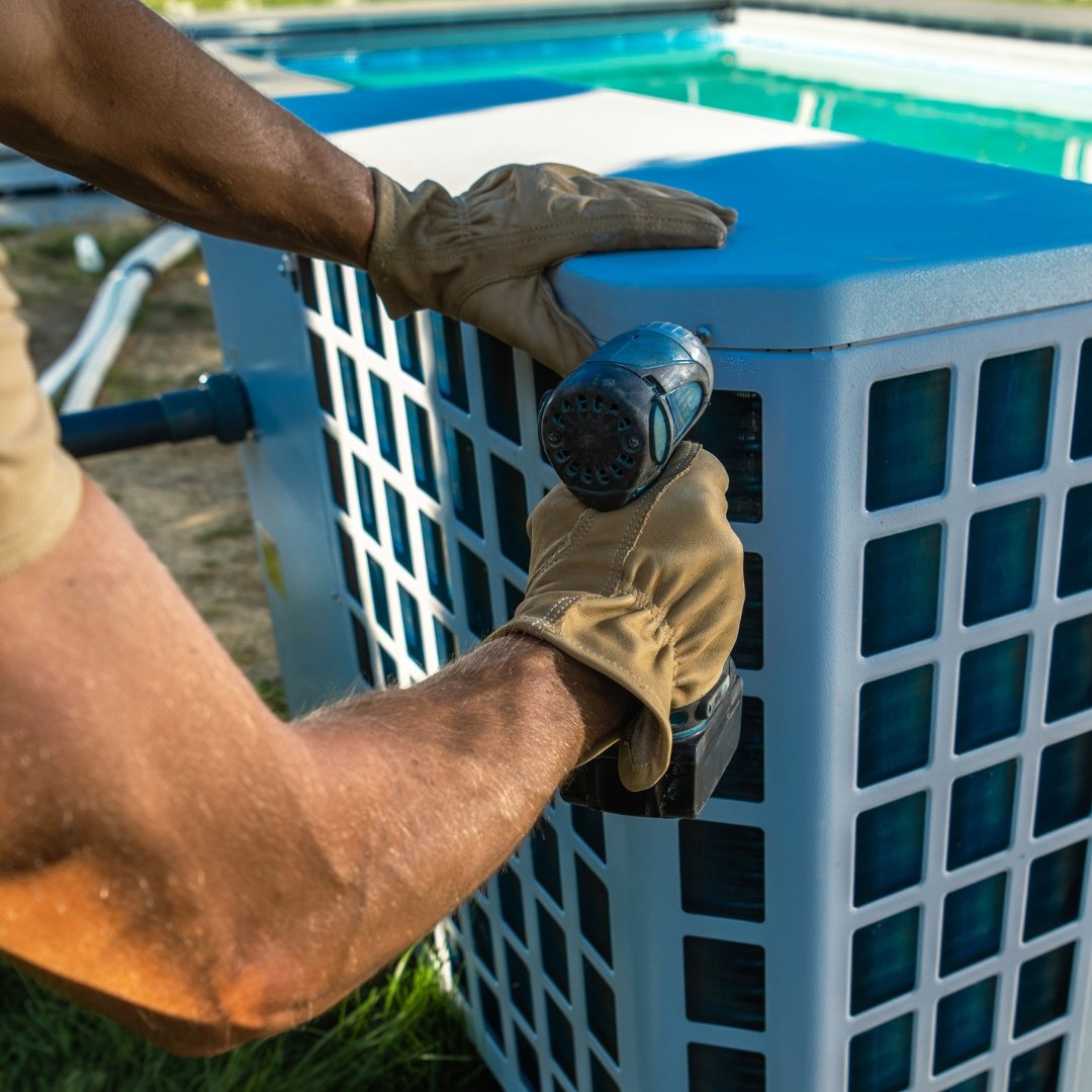 Person wearing gloves using a cordless drill to secure a panel on a large outdoor air conditioning or refrigeration unit with pool in background.