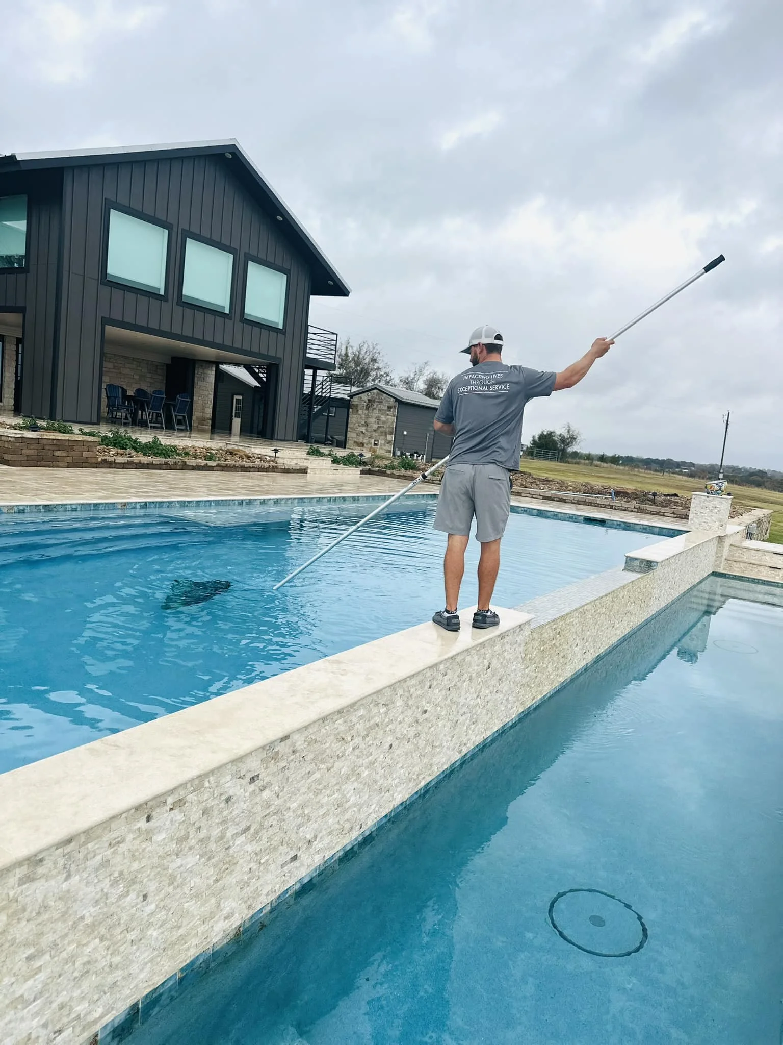 A man standing on the edge of a swimming pool, using a long pole to clean the pool.
