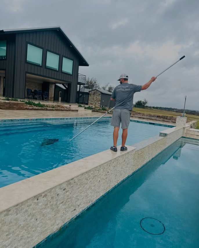 Person cleaning a swimming pool with a pool skimmer, standing on the edge of the pool