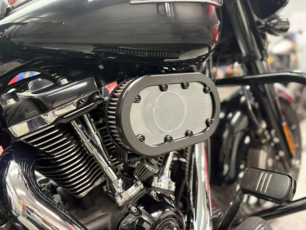 Close-up of a motorcycle engine with a cover removed, showing the chrome finish and black fins, with surrounding components and a black fuel tank.