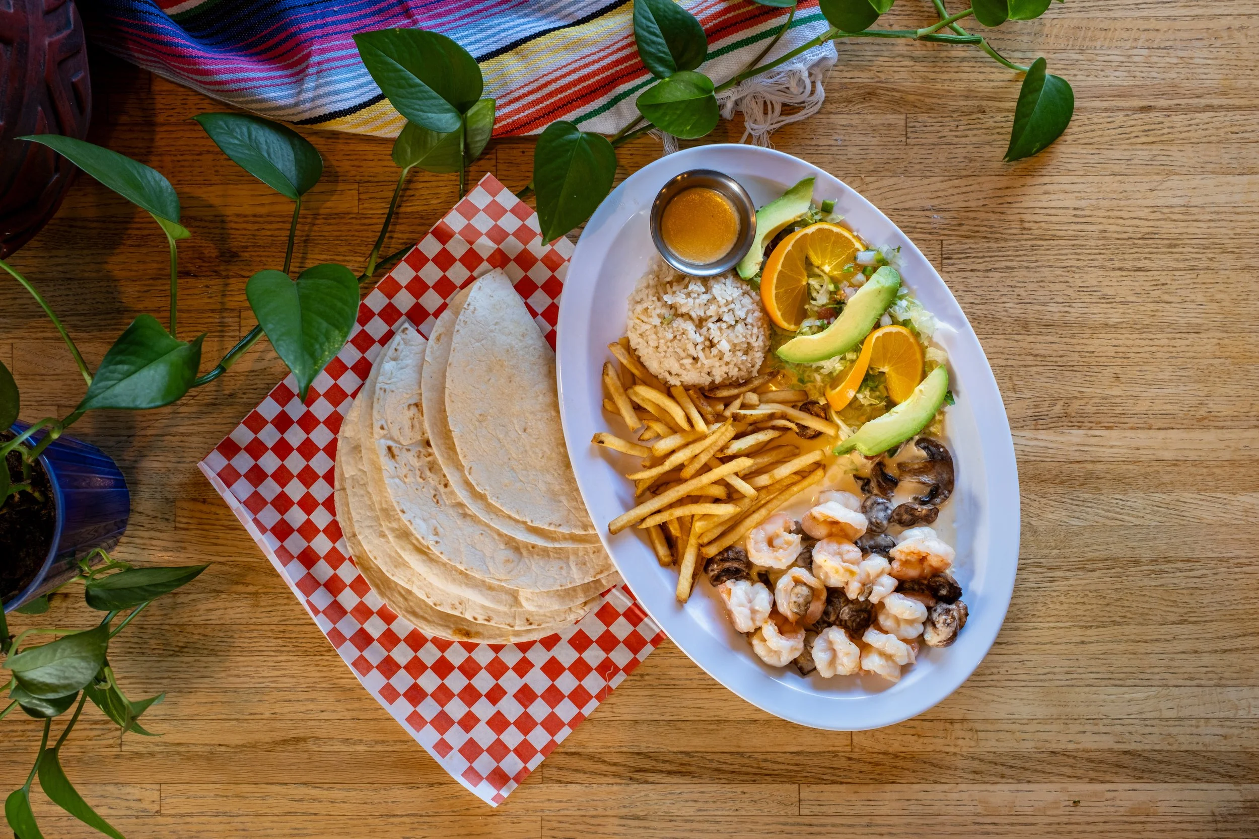 A Mexican seafood dish served on an oval white plate with rice, french fries, avocado slices, orange slices, and a small container of sauce. Accompanying the plate are three tortillas on a red and white checkered paper, all set on a wooden table with