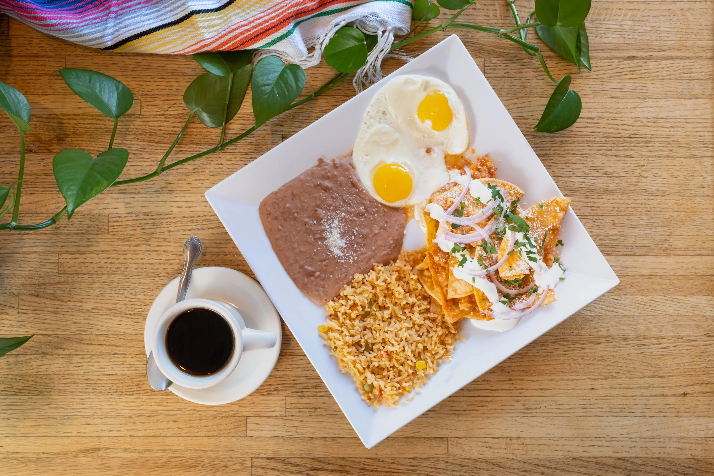 Mexican breakfast plate with two sunny side up eggs, refried beans, rice, and nachos with sour cream and cilantro, alongside a cup of black coffee on a wooden table with a green plant and a striped cloth nearby.
