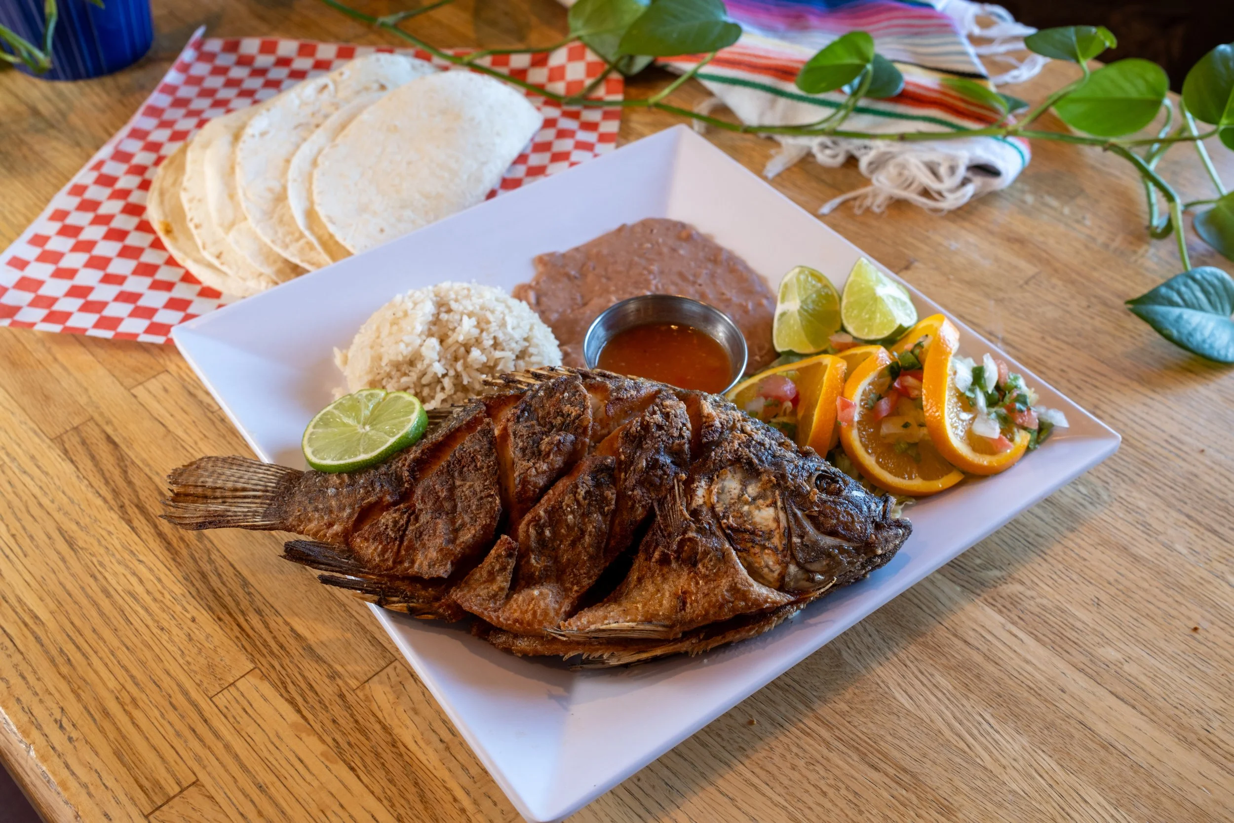 A plate of grilled fish with lime wedges, rice, a side of beans, salsa, and sliced citrus fruits. Behind the plate, there are tortillas on checkered paper, with a cloth napkin and green plant leaves on a wooden table.