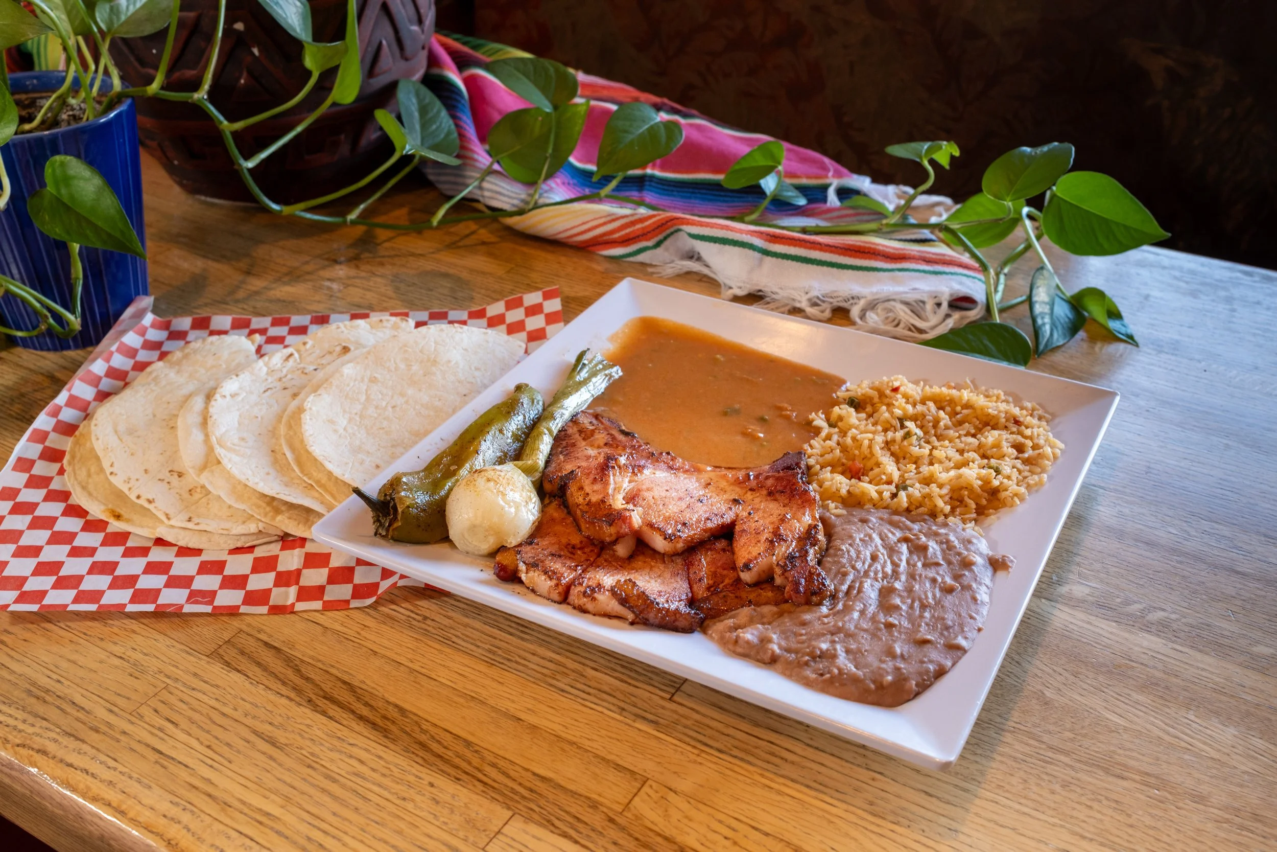 A Mexican meal on a wooden table with tortillas, rice, beans, grilled meat, green chili pepper, onion, and sauce, with a colorful cloth and potted plant in the background.