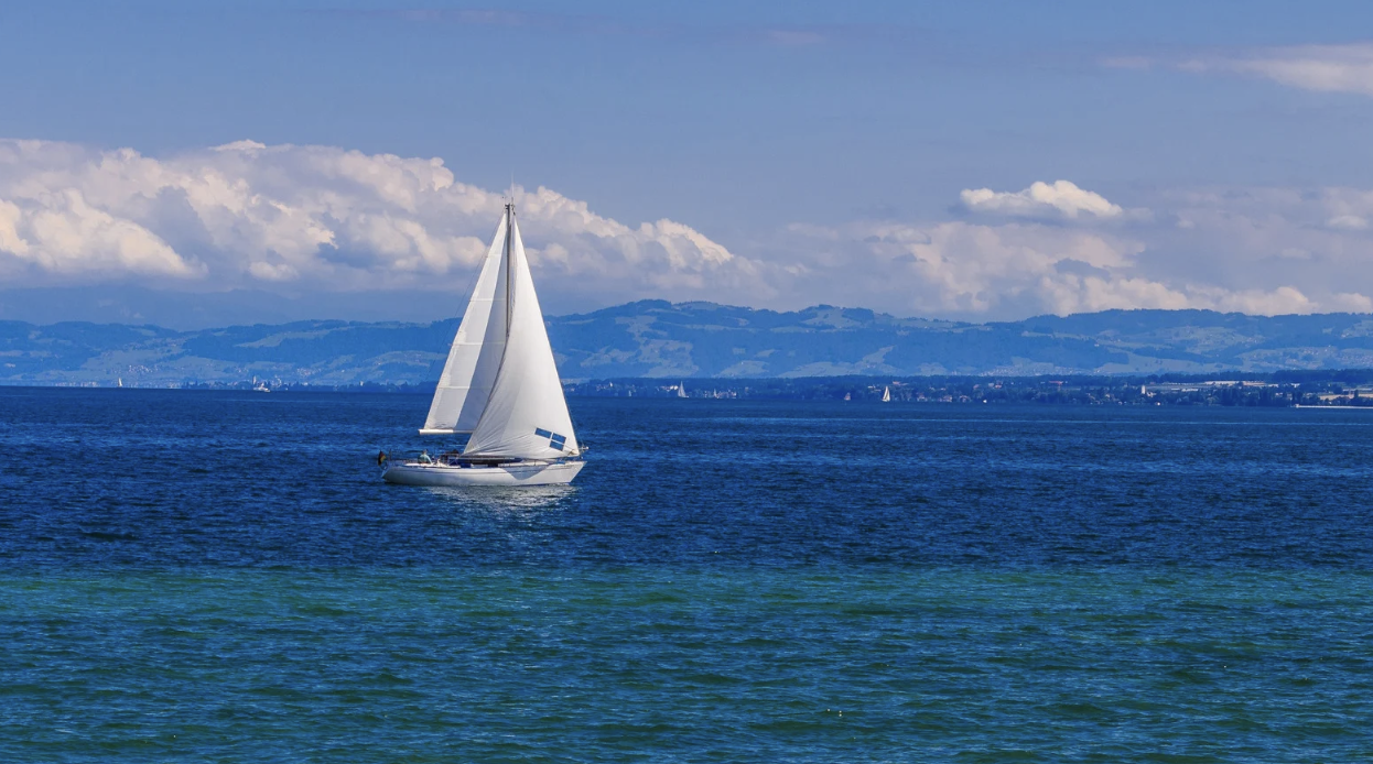 Segelboot auf einem blauen See mit Bergen im Hintergrund und einer bewölkten Himmelsszene.