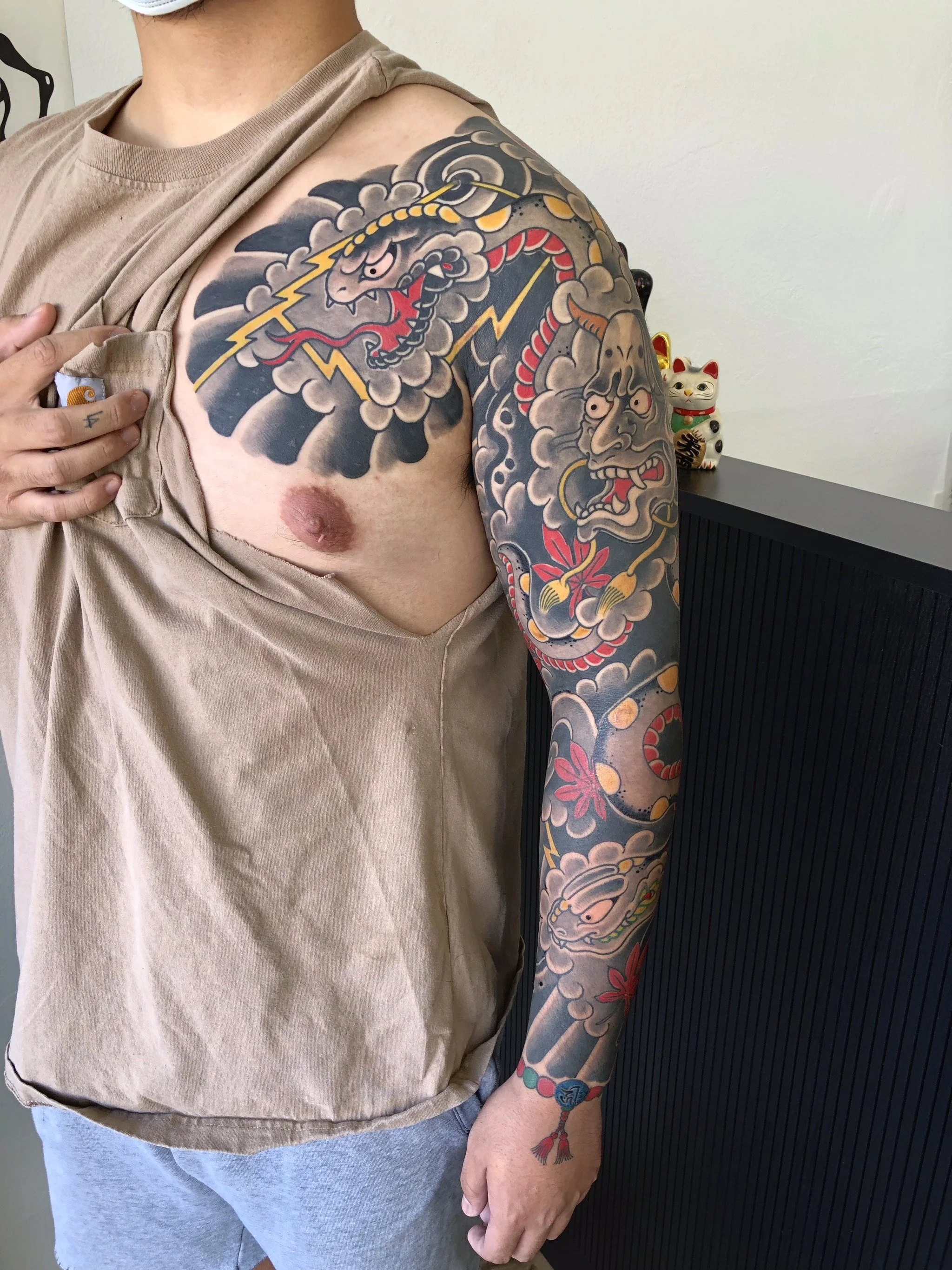 A person with a shoulder-length sleeve tattoo featuring a dragon, clouds, lightning bolts, a face, and flowers in traditional Japanese style, wearing a beige shirt and gray shorts.
