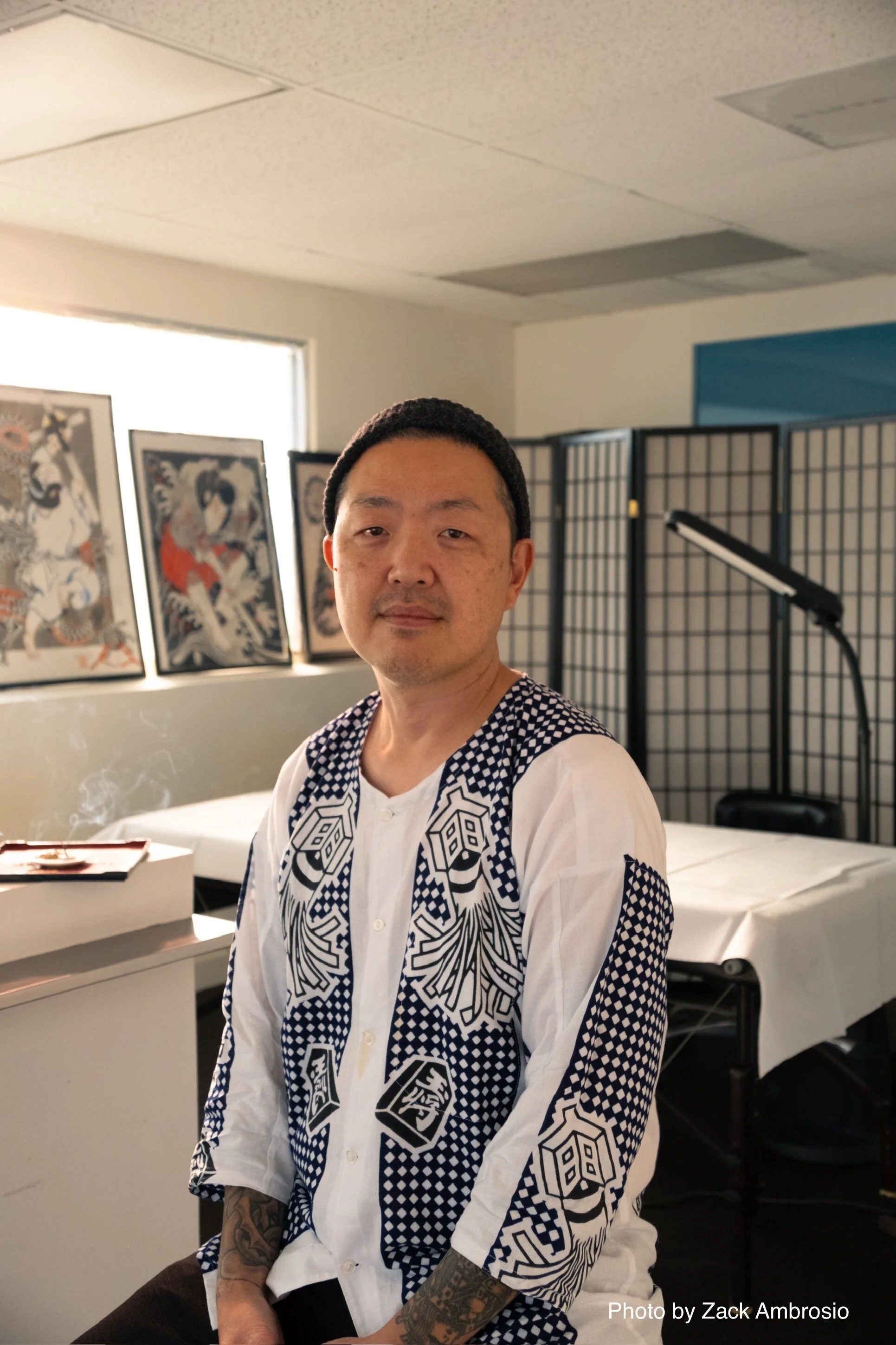 A man with tattoos on his hands and a black beanie, sitting in an indoor space with artwork on the wall behind him and a white table nearby.