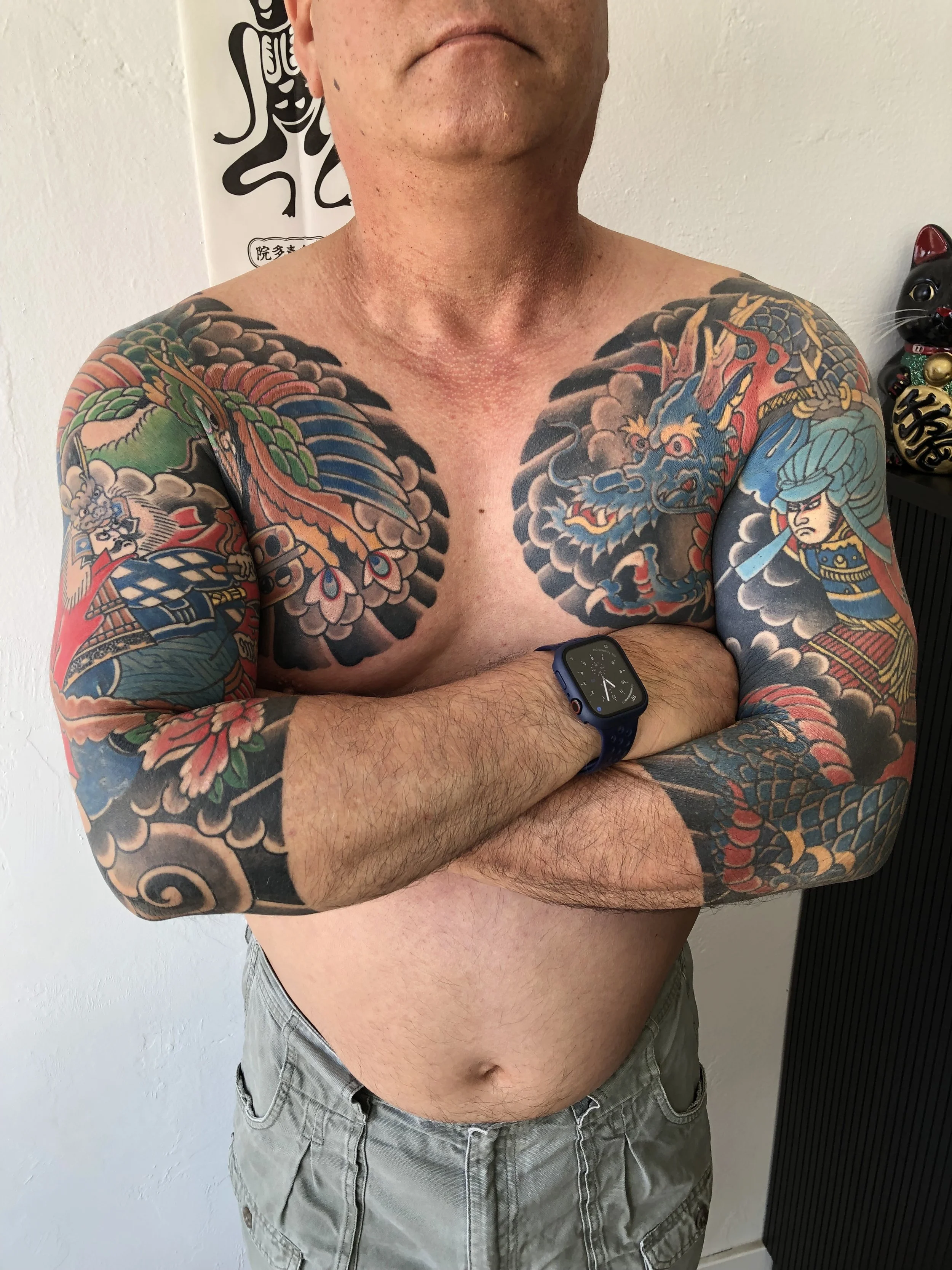 A shirtless man with colorful Japanese-style tattoos across his chest and arms, wearing a blue smartwatch, standing with arms crossed in front of a white wall.
