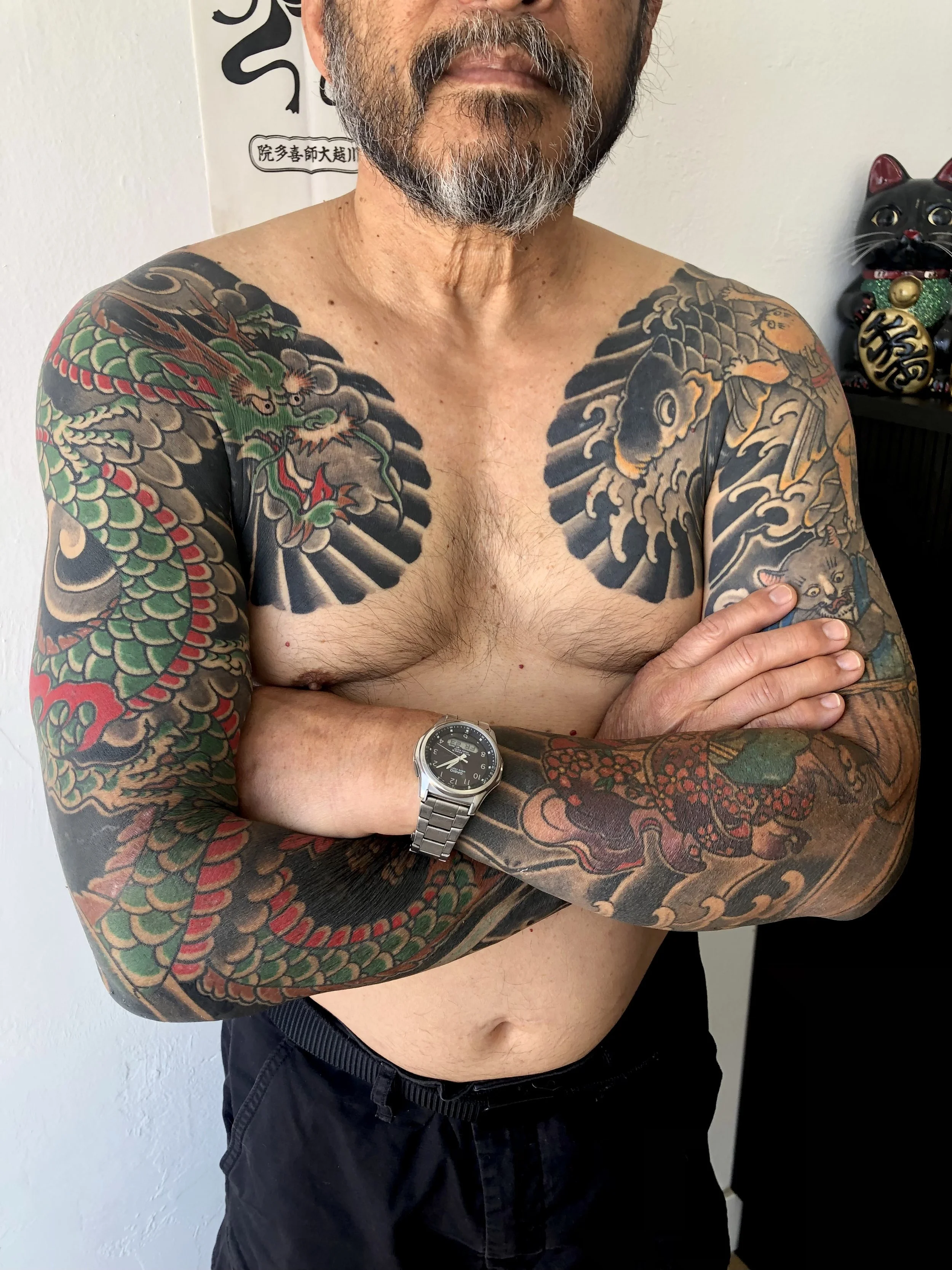 A shirtless man with tattoos on his chest and arms, crossing his arms, wearing a silver watch, and standing indoors with Japanese artwork and a lucky cat figurine in the background.
