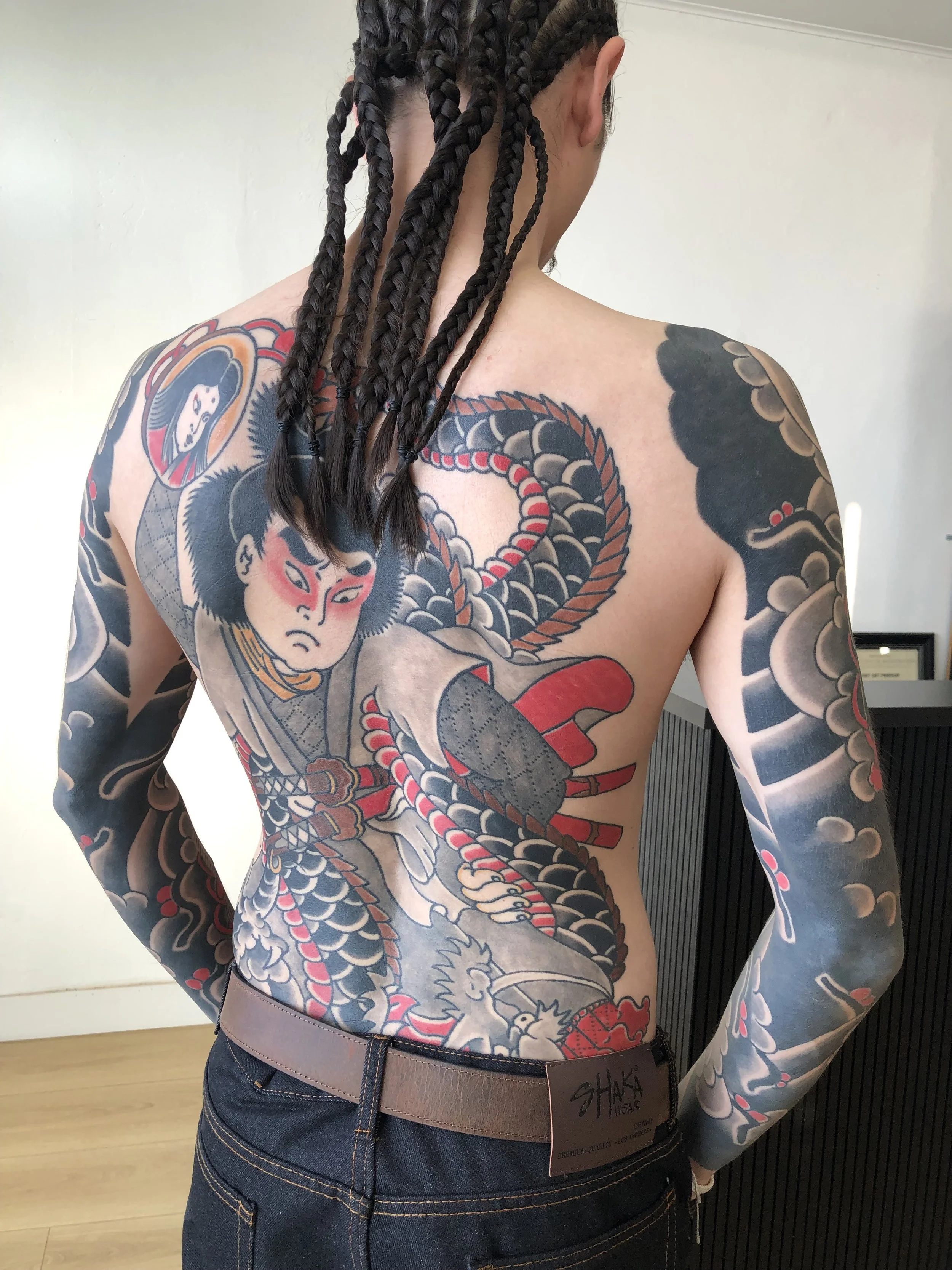 A person with elaborate Japanese-style body tattoos featuring a samurai, a dragon, and a woman, with long braided hair and wearing dark jeans with a brown belt.