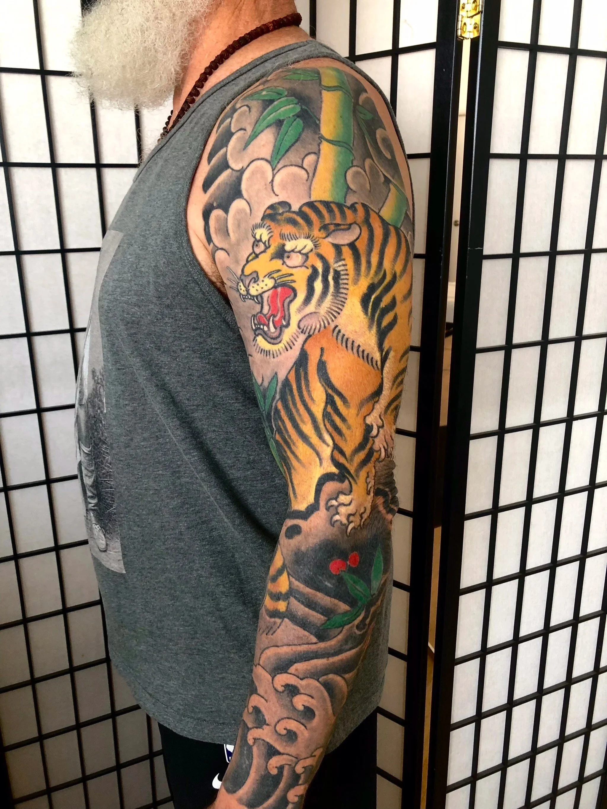 Tattoo of a roaring tiger on a person's upper arm and shoulder, with bamboo and clouds in the background.