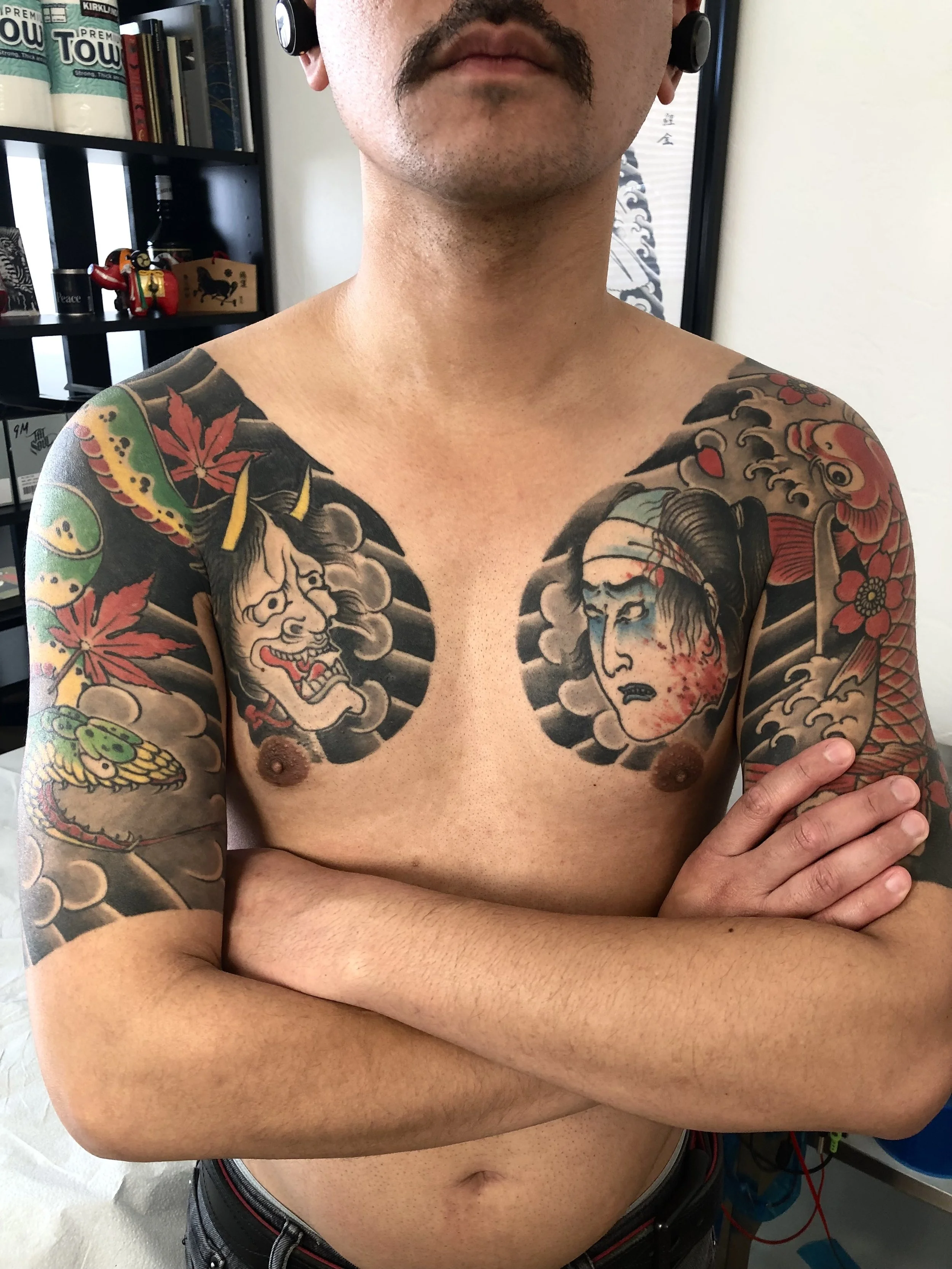 A shirtless man with tattoos on his chest and arms, standing with arms crossed in a room with shelves and artwork in the background.