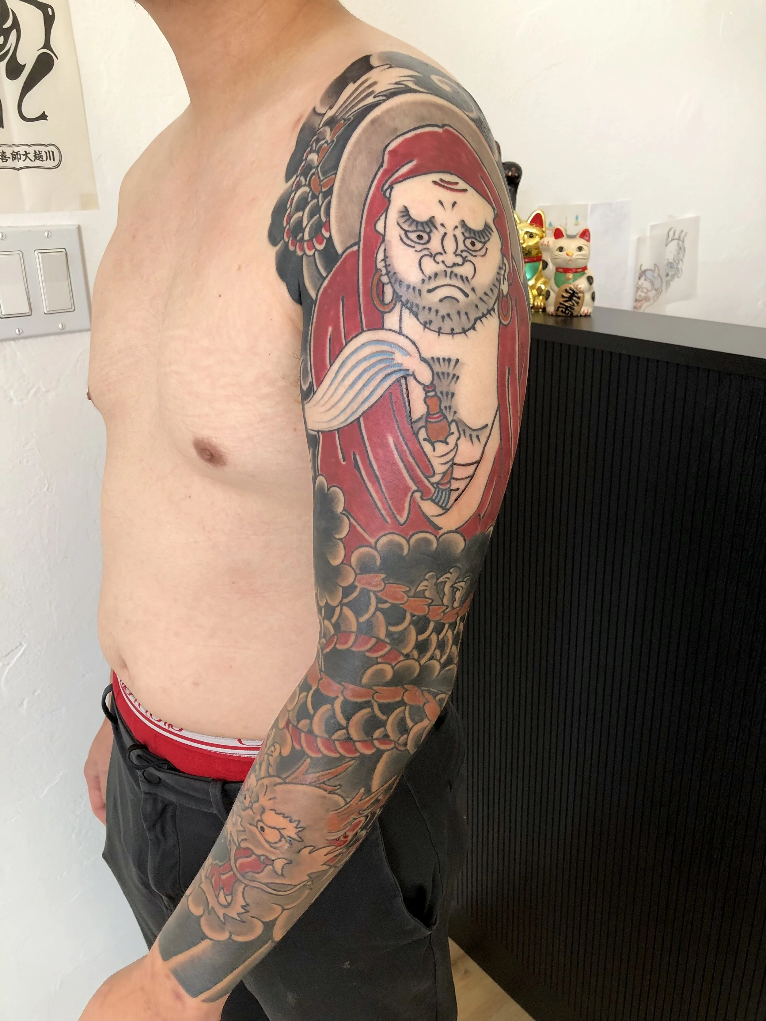 Tattoo of a traditional Japanese warrior on upper arm, with a fierce face, red hood, and holding a sword, surrounded by clouds and a coiled dragon.