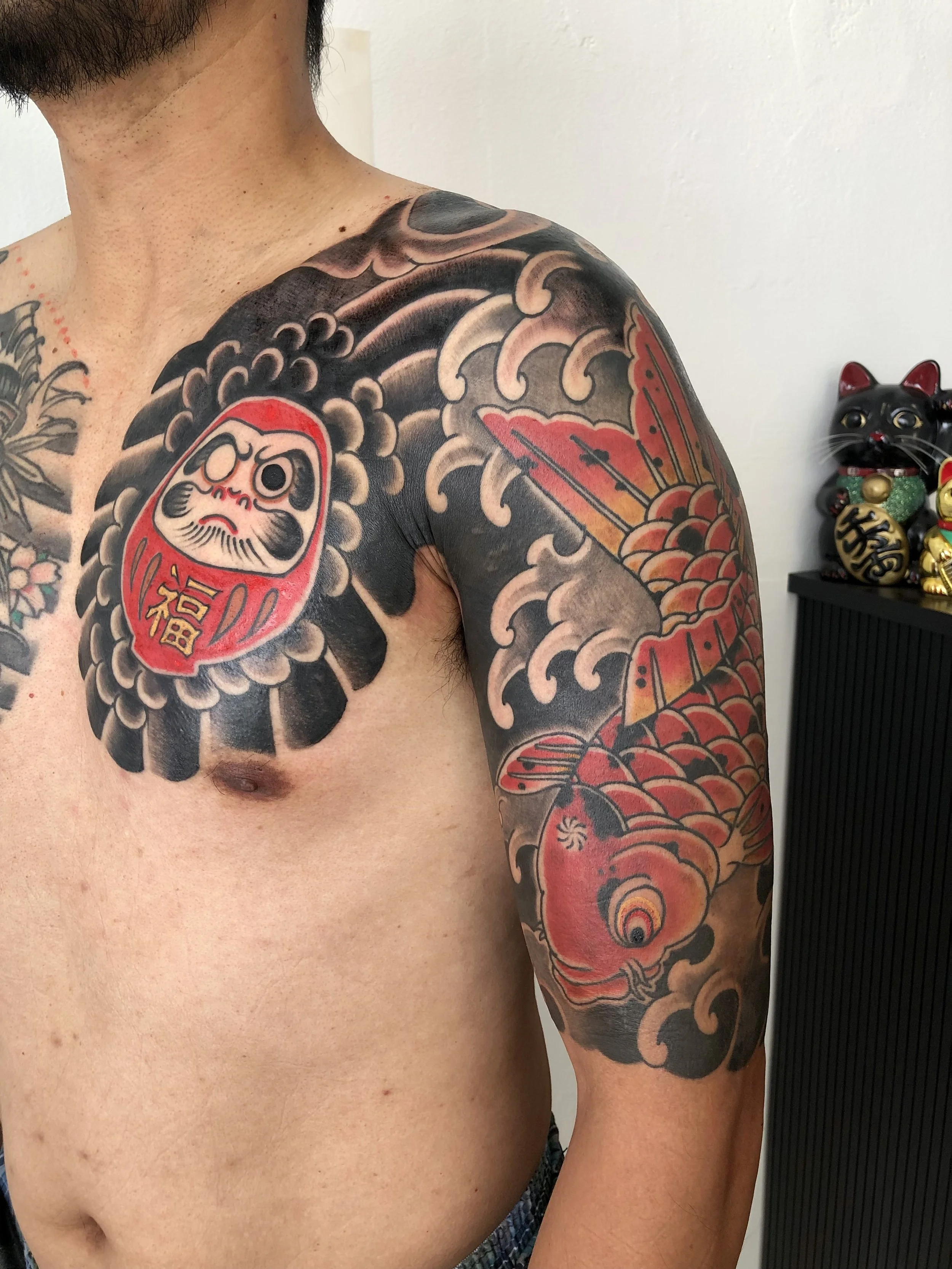 Tattoo of a red Daruma doll on the chest, with black and red traditional Japanese wave and fish (koi) designs on the shoulder and arm.