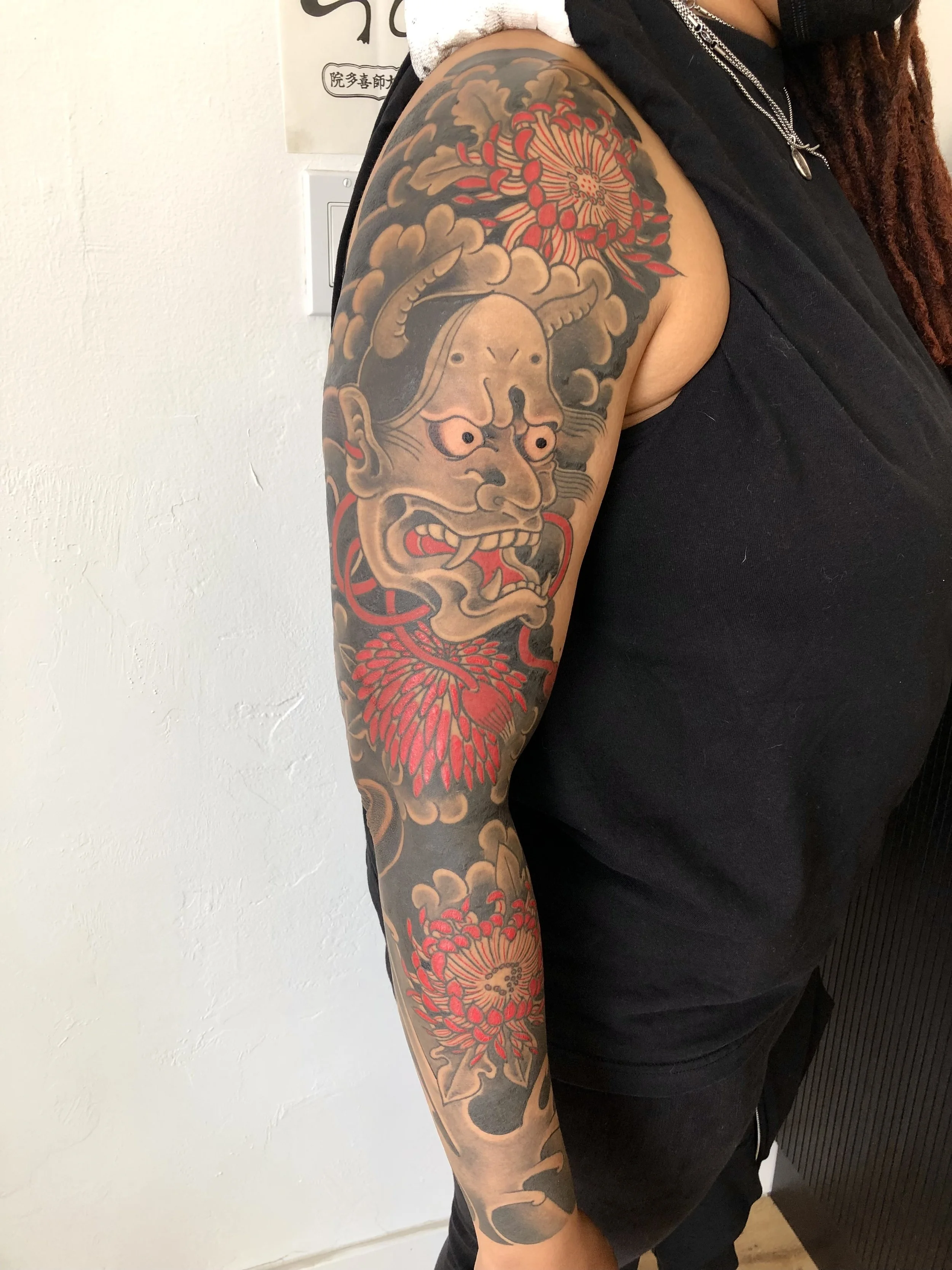 A person with a full sleeve tattoo on their left arm featuring a Japanese-style Hannya mask with a fierce expression, surrounded by red and black floral and cloud designs.