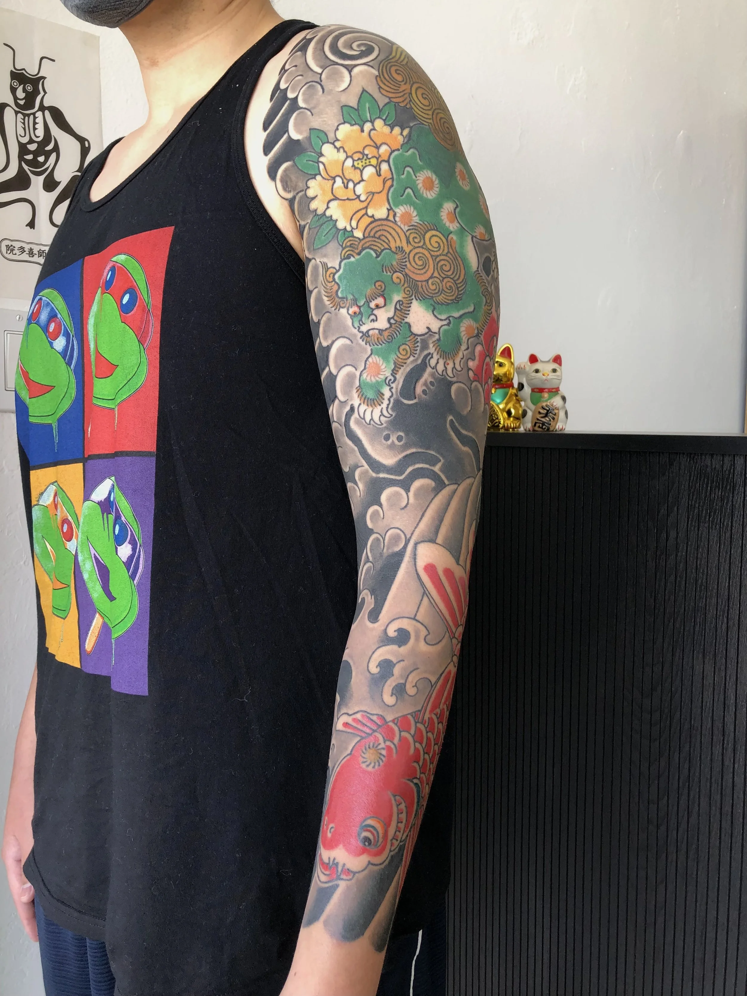 A person with a colorful sleeve tattoo featuring traditional Japanese-style imagery, including a lion, clouds, and a koi fish, wearing a black tank top with a colorful pop art design.