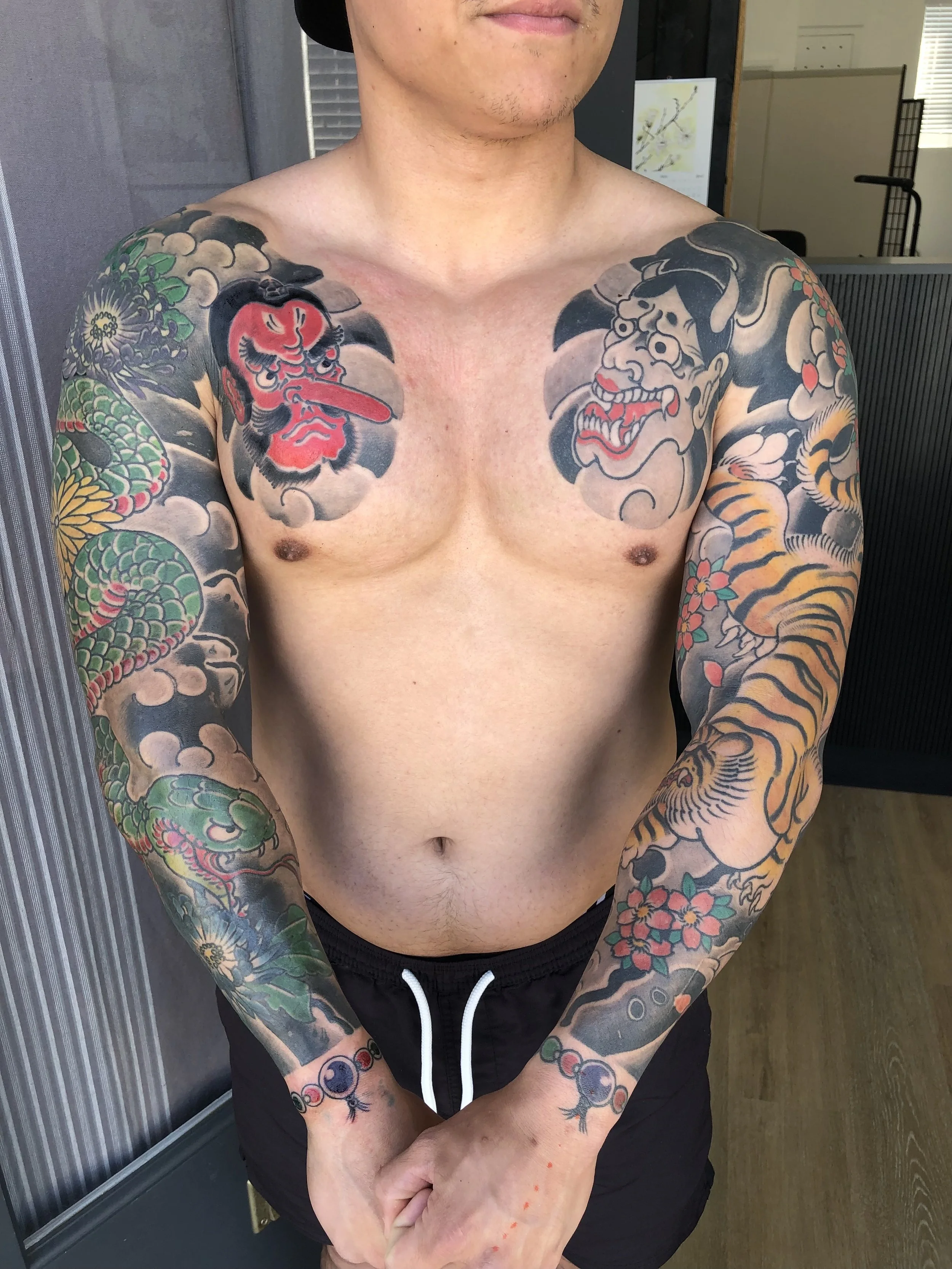 A shirtless man with tattoos on his arms and chest, featuring traditional Japanese-style mythological characters and designs, standing indoors with arms crossed in front of him.