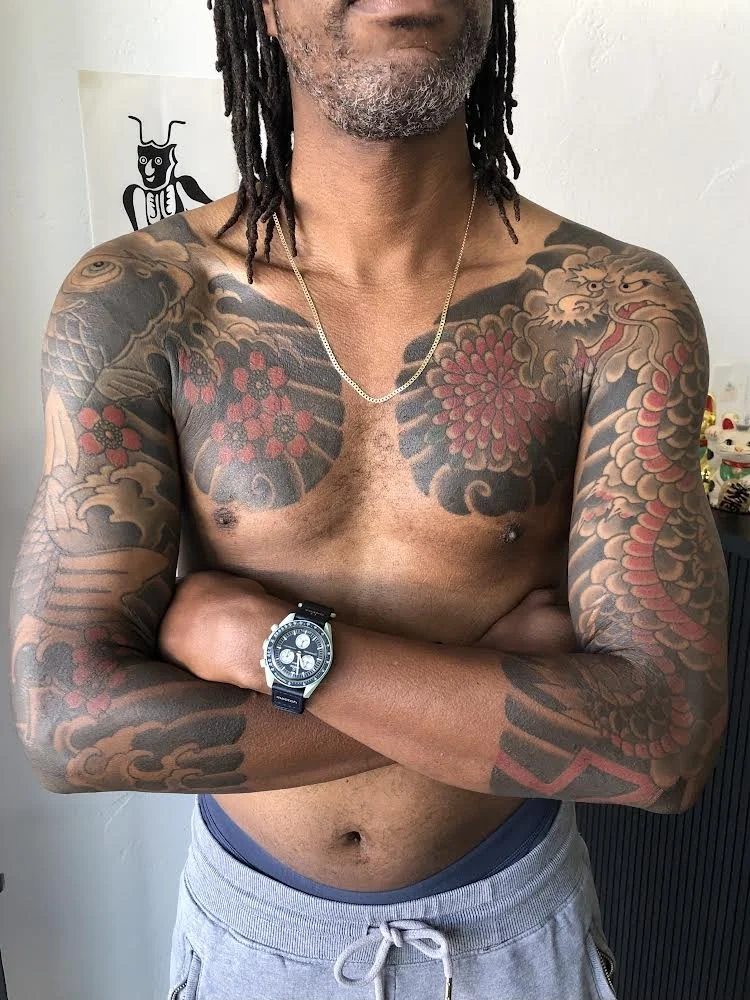 A shirtless man with tattoos on his chest and arms, wearing a gold chain and black watch, crossing his arms in front of a white wall with a cartoon drawing of a dog.