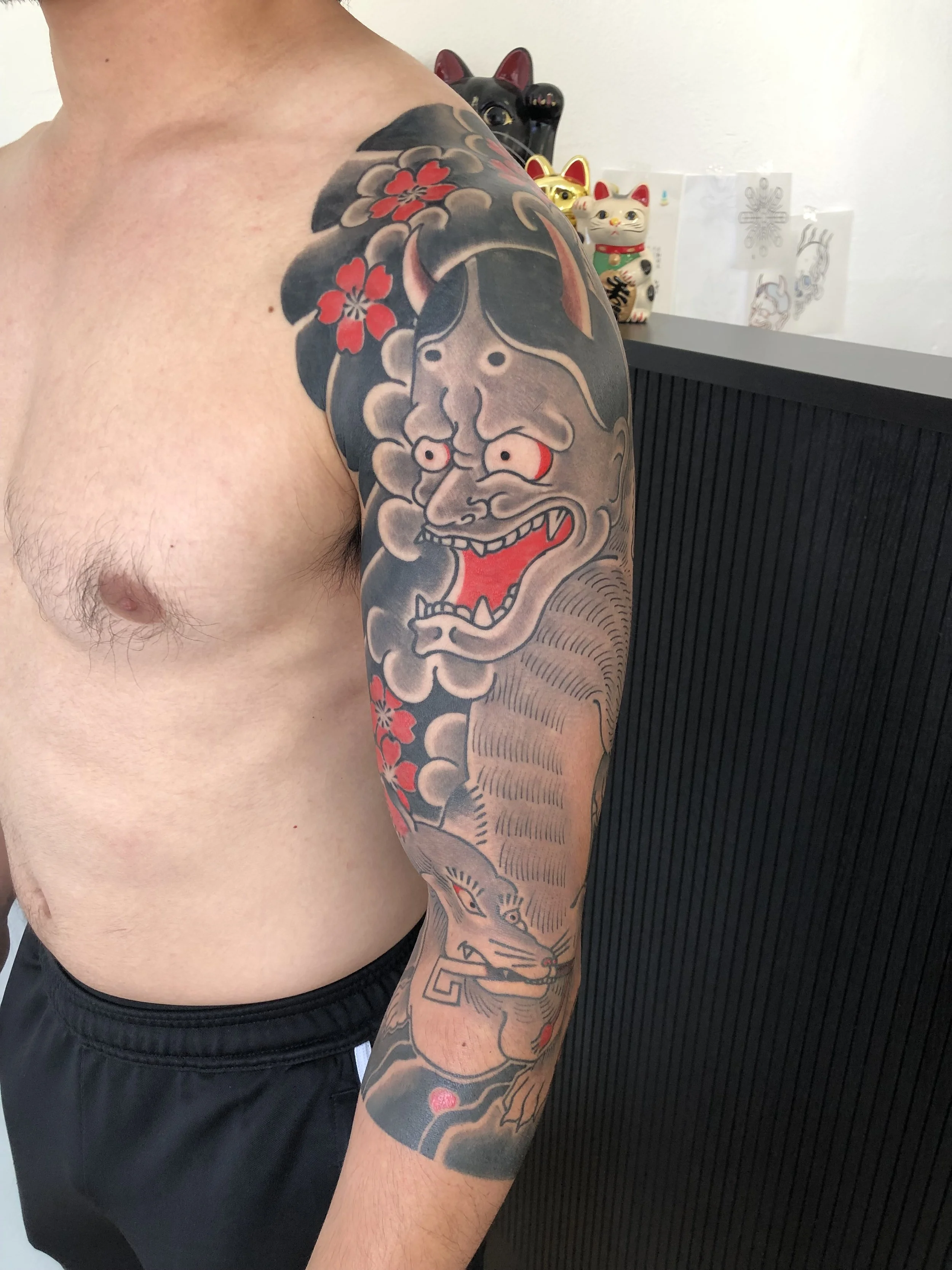 Tattoo of a fierce demon face and a lunar tiger on a man's upper arm, with cherry blossoms and clouds in traditional Japanese style.