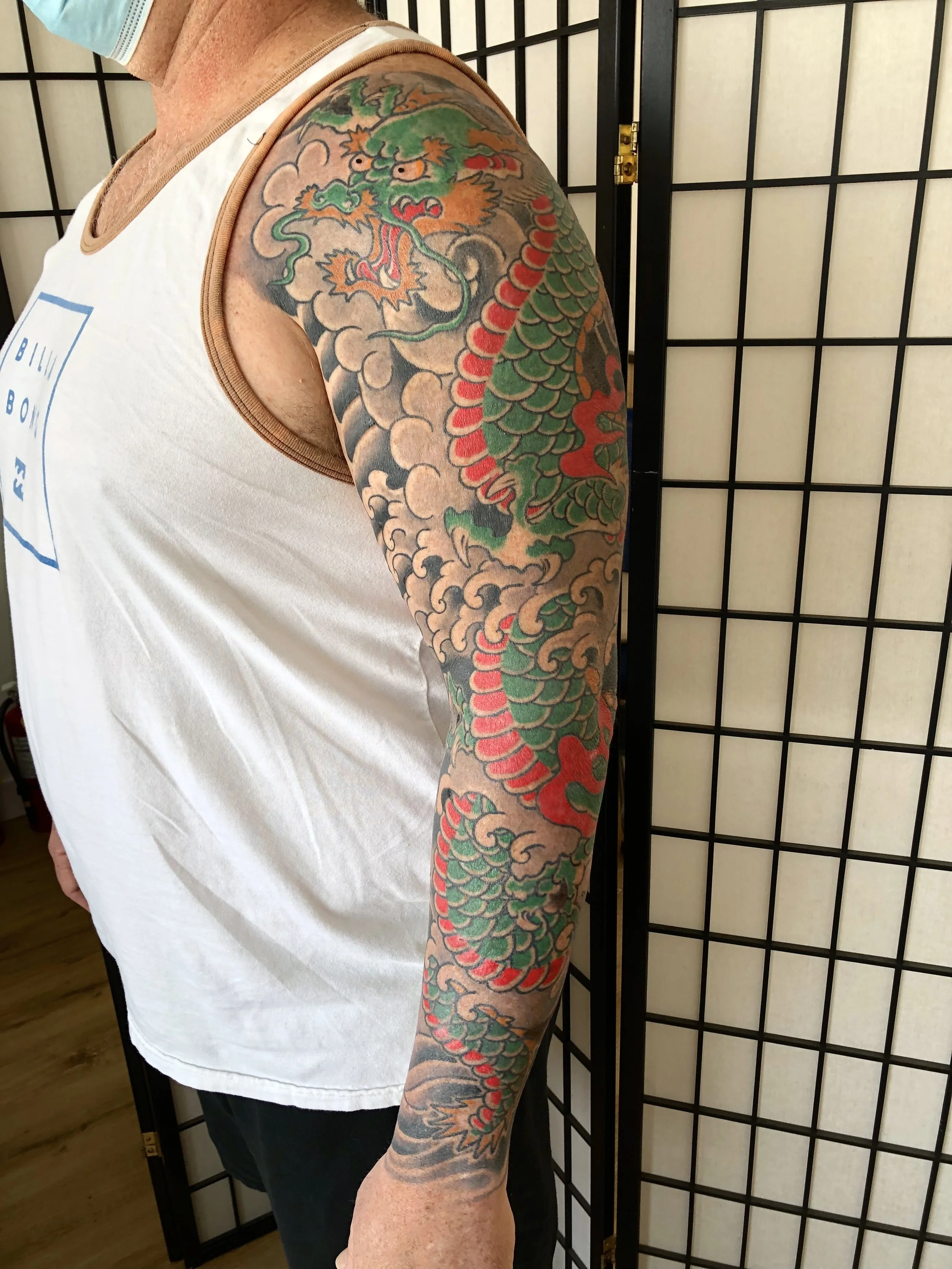 A person with a large colorful dragon tattoo on their arm, featuring green, red, and gold scales, set against a background of clouds. The person is wearing a white sleeveless shirt and a face mask, standing in front of a black folding screen.