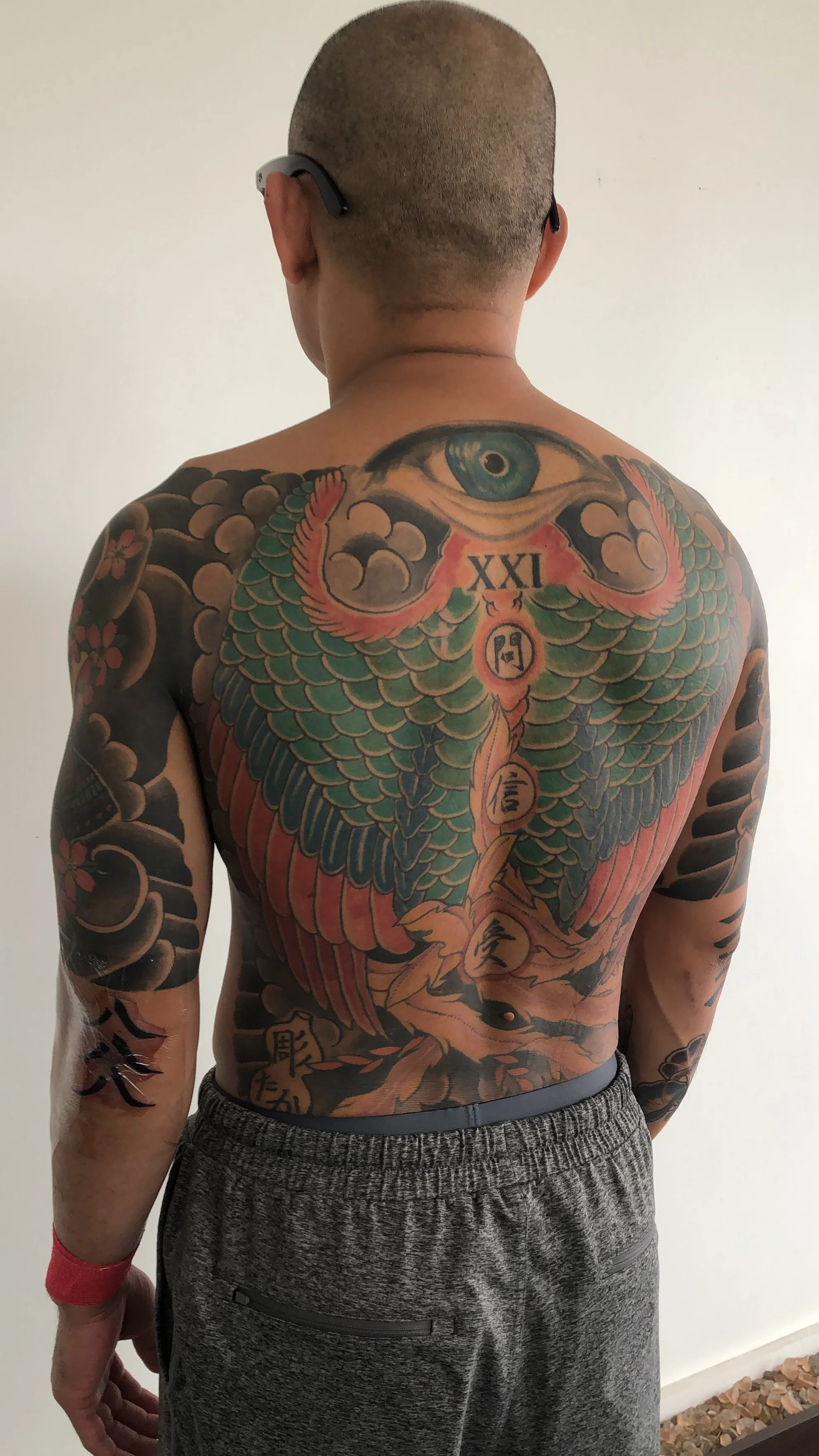 Back view of a man with extensive colorful tattoos, including a large owl and eye on his back, and Japanese characters on his arm.